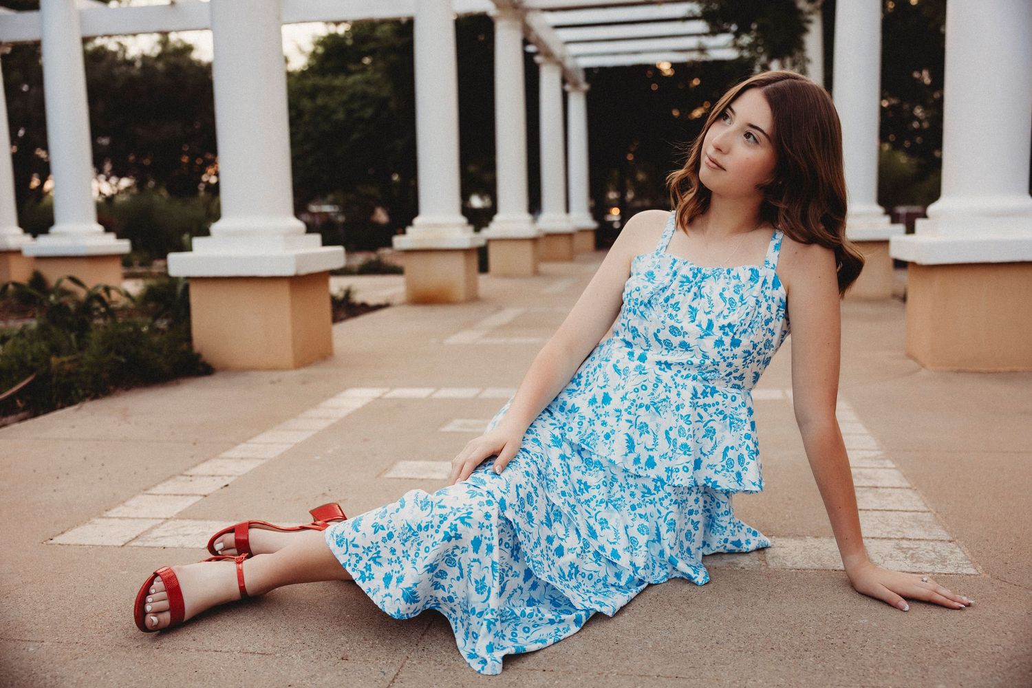 Abilene High School Senior in blue floral midi dress and red sandals sitting gracefully on tiled patio with white columns.