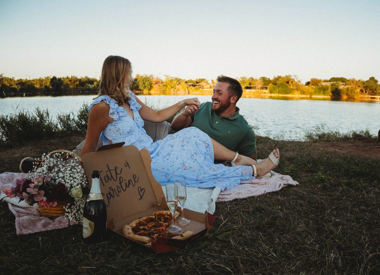 Engagement photo session by a lake in Abilene, Texas featuring a couple sitting on a picnic blanket during golden hour.