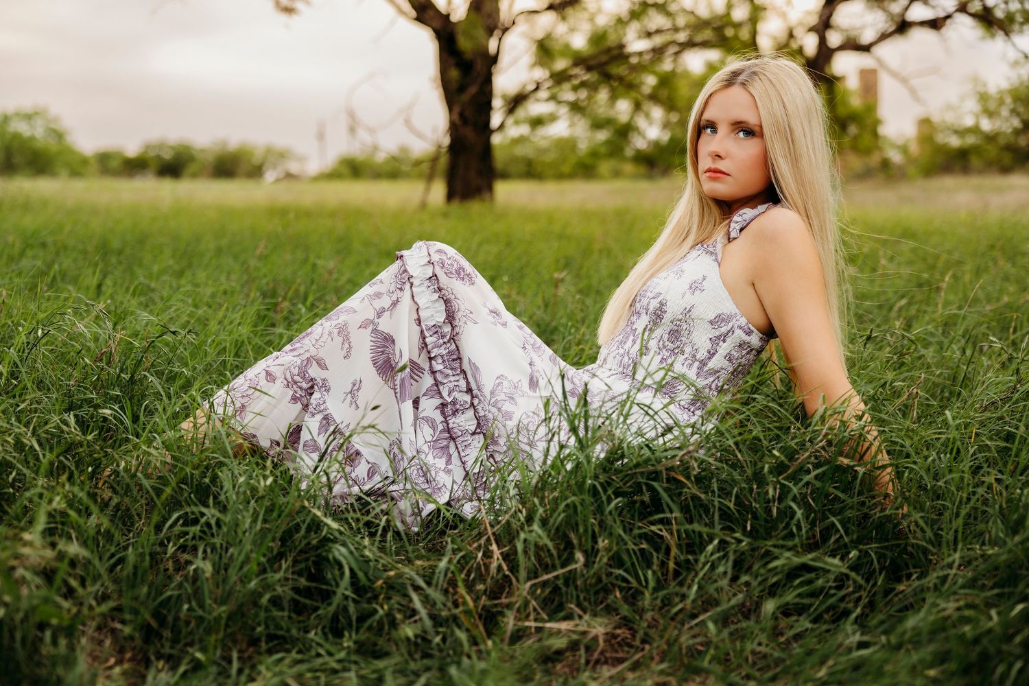 Blonde high school senior in a floral dress sitting in an open field during outdoor senior photos in Abilene, TX.