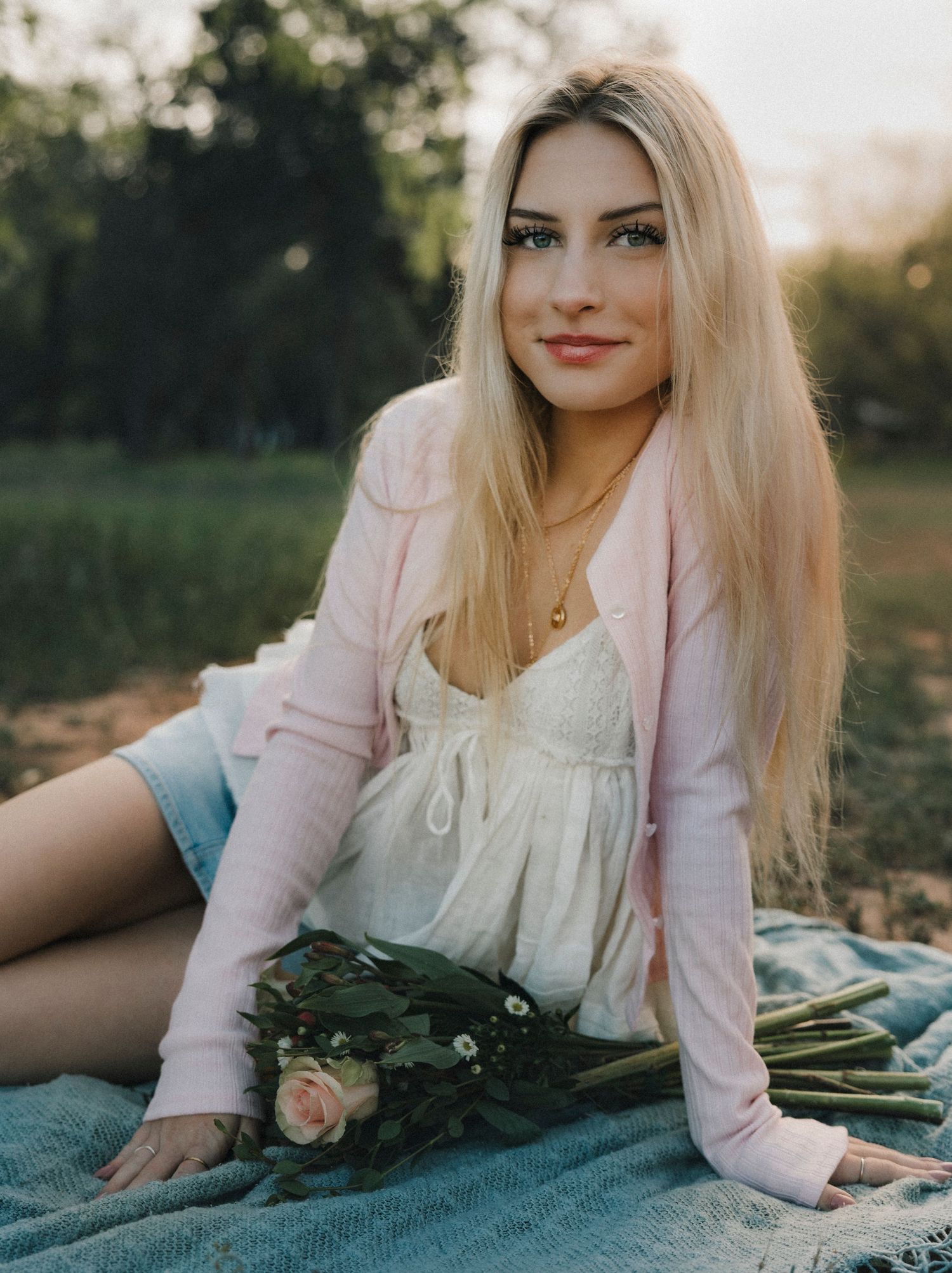 Senior photoshoot in pink cardigan and white top sits on blanket outdoors holding roses, smiling softly at camera.