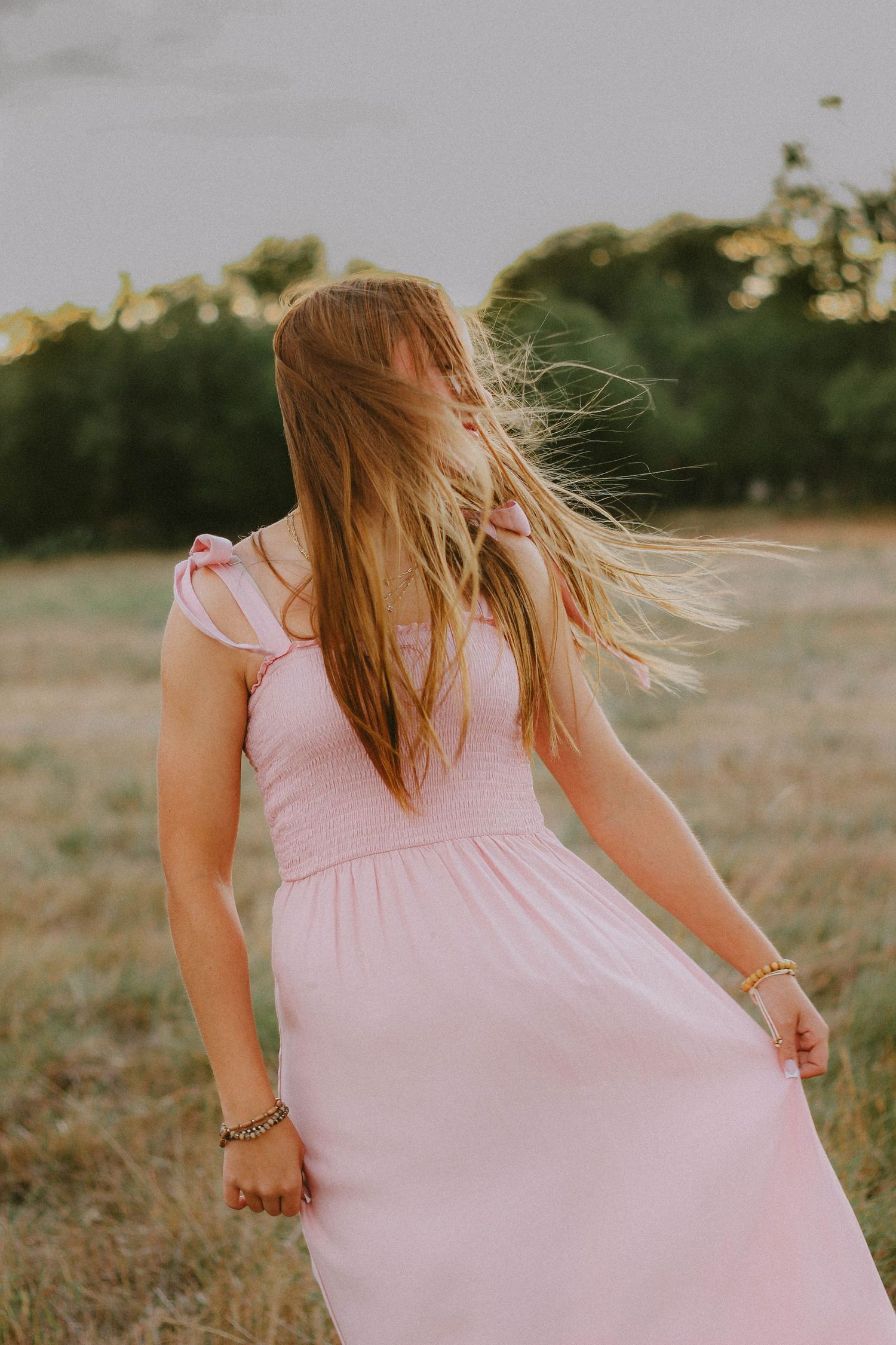 Senior photoshoot in pink dress with hair blowing in wind standing in golden hour field.