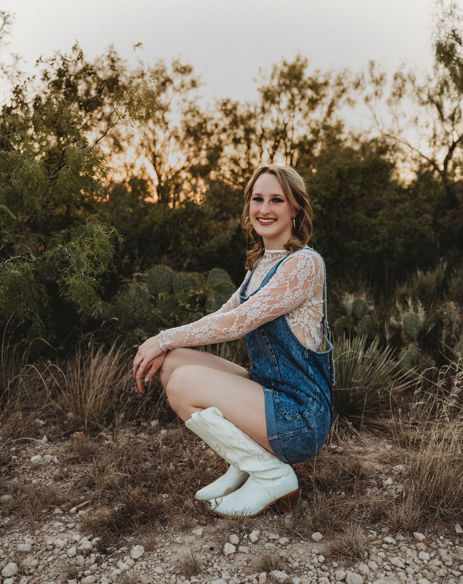 Smiling Class of 2027 senior crouching among native Texas grasses during a natural light senior portrait session in Abilene, Texas.