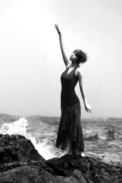 Black and white senior portrait of high school senior standing on rocks by the waterfall in a flowing dress