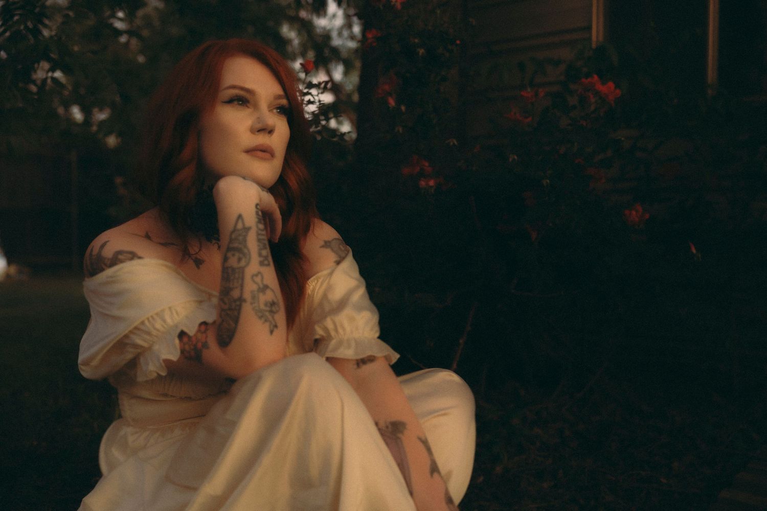 Tattooed redhead woman in white off-shoulder dress sitting outdoors at dusk, gazing thoughtfully.