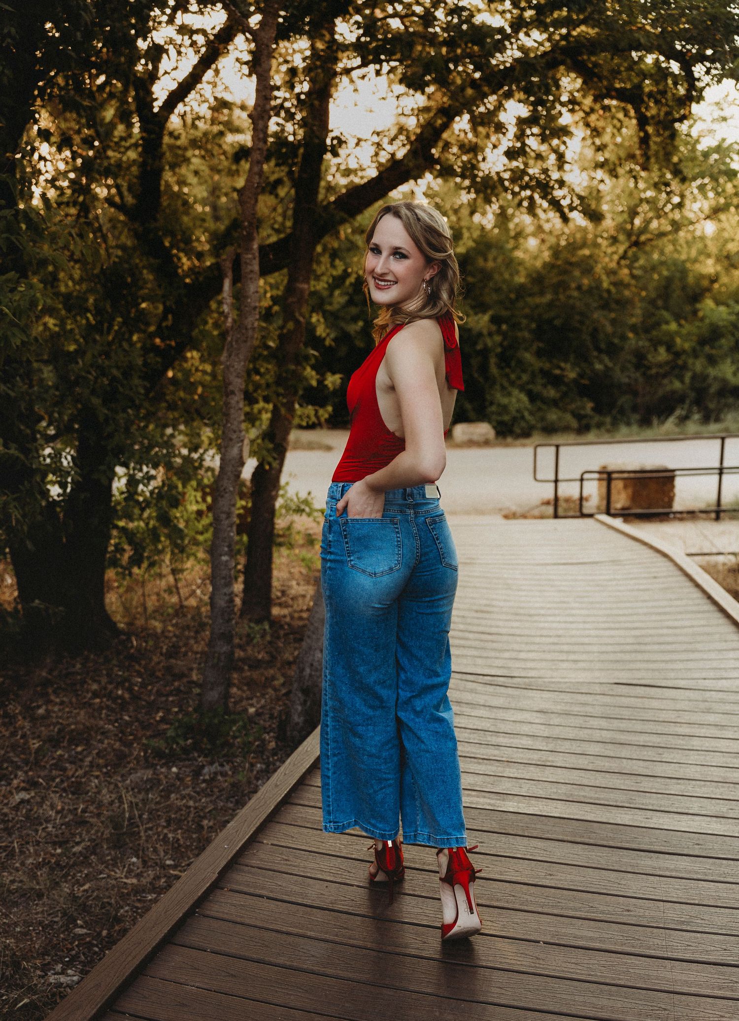 High school senior walking across a wooden bridge in a red top and jeans during a golden hour senior photography session in Abilene, Texas.