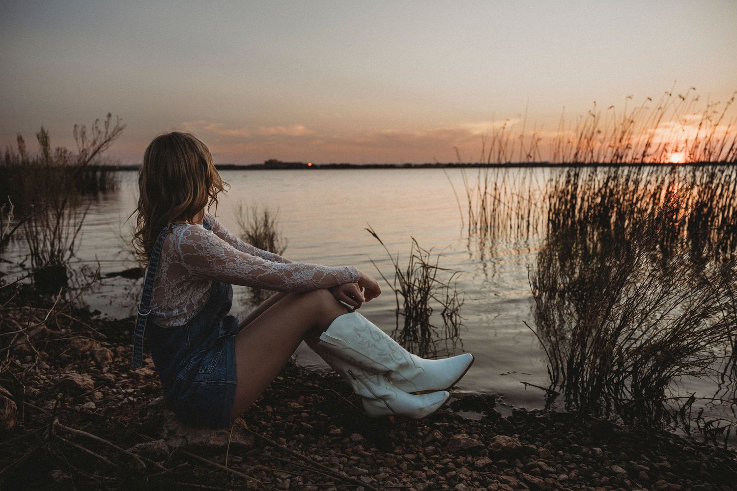 High school senior sitting along the shoreline watching the sunset during a lakeside senior photography session in Abilene, Texas.