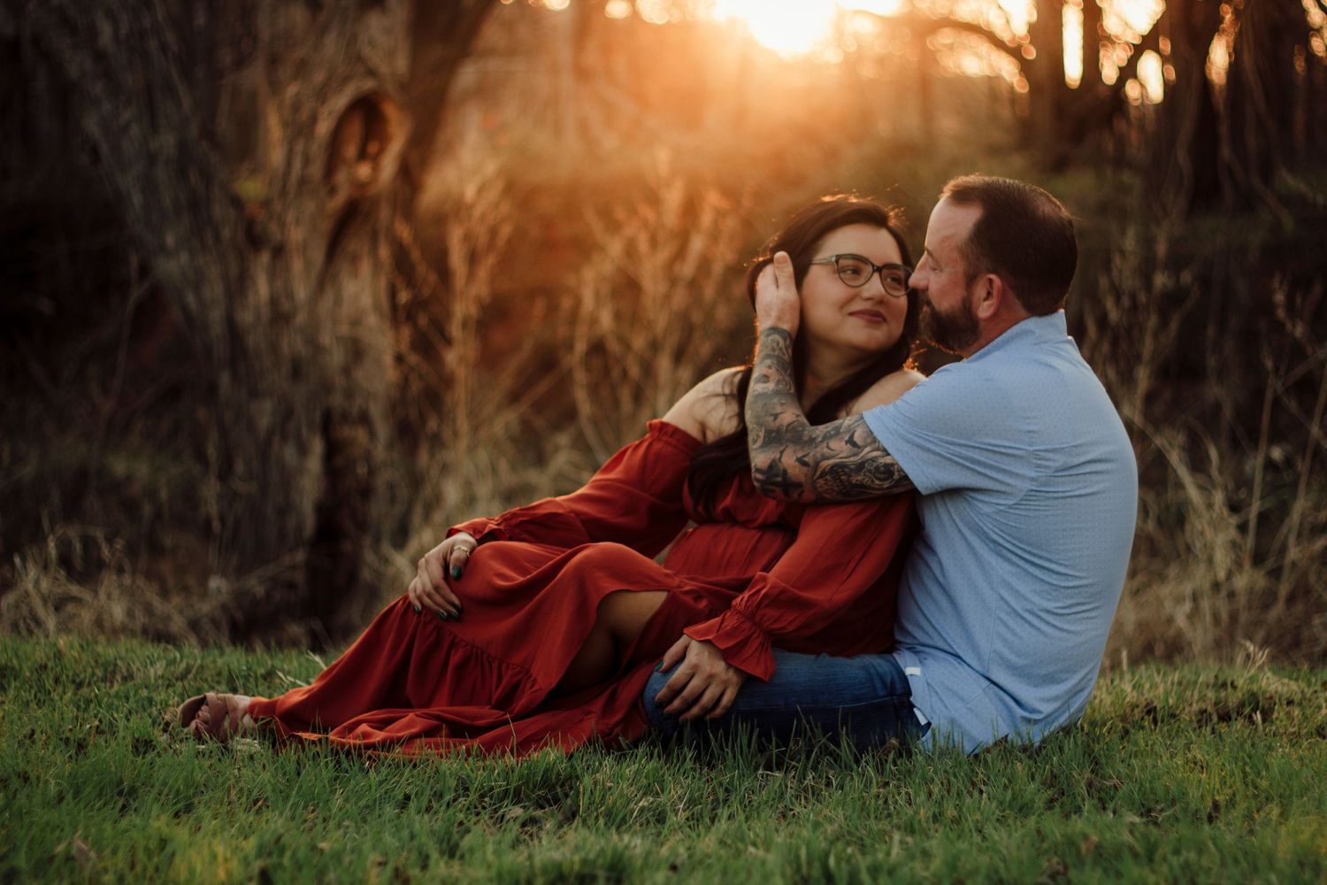 Couple sitting on grass at sunset, woman in red dress leaning against tattooed man in blue shirt, golden hour light.