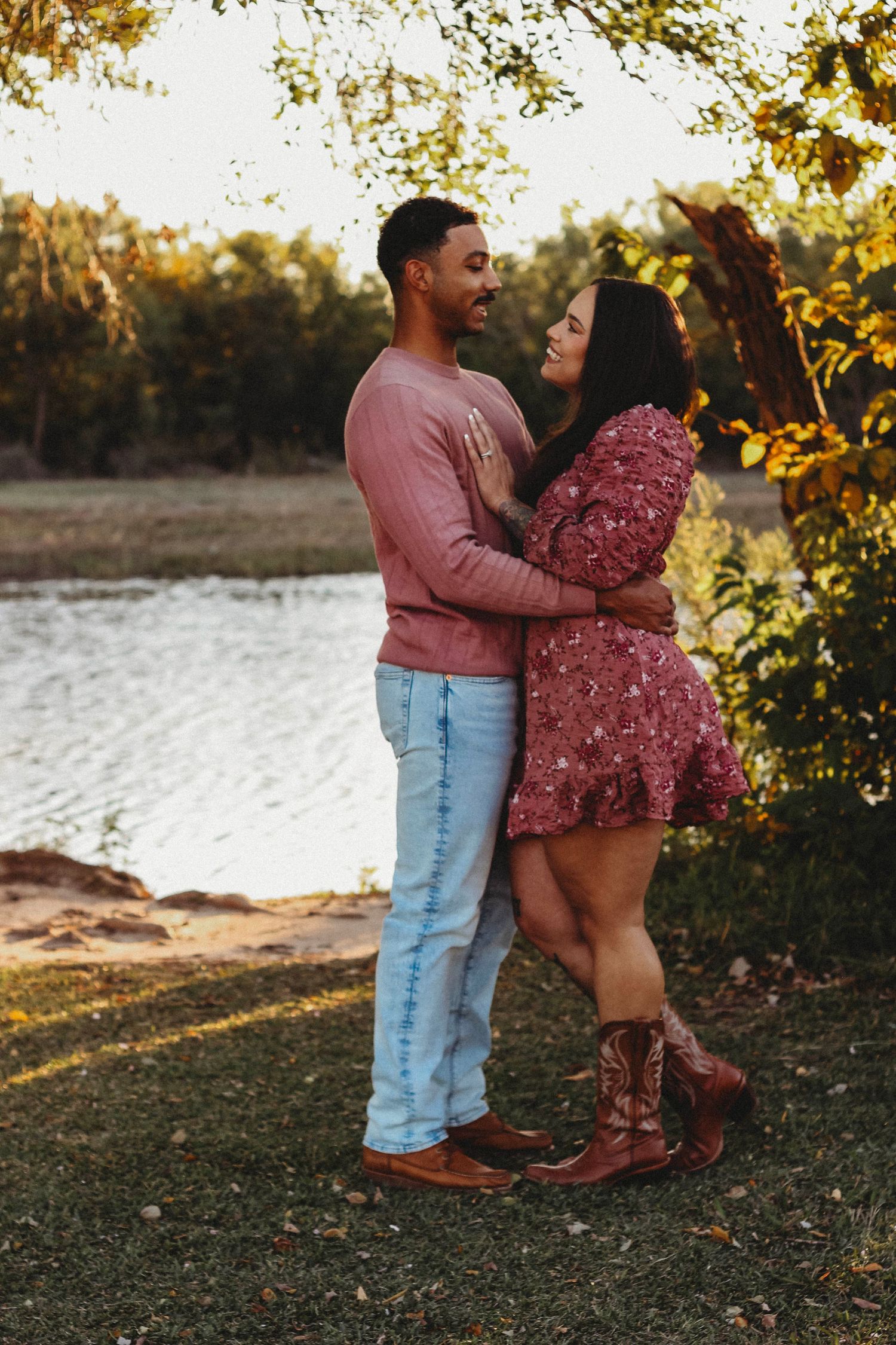 Couple embracing near a lake at golden hour, woman in floral dress and cowboy boots, man in pink sweater and jeans.
