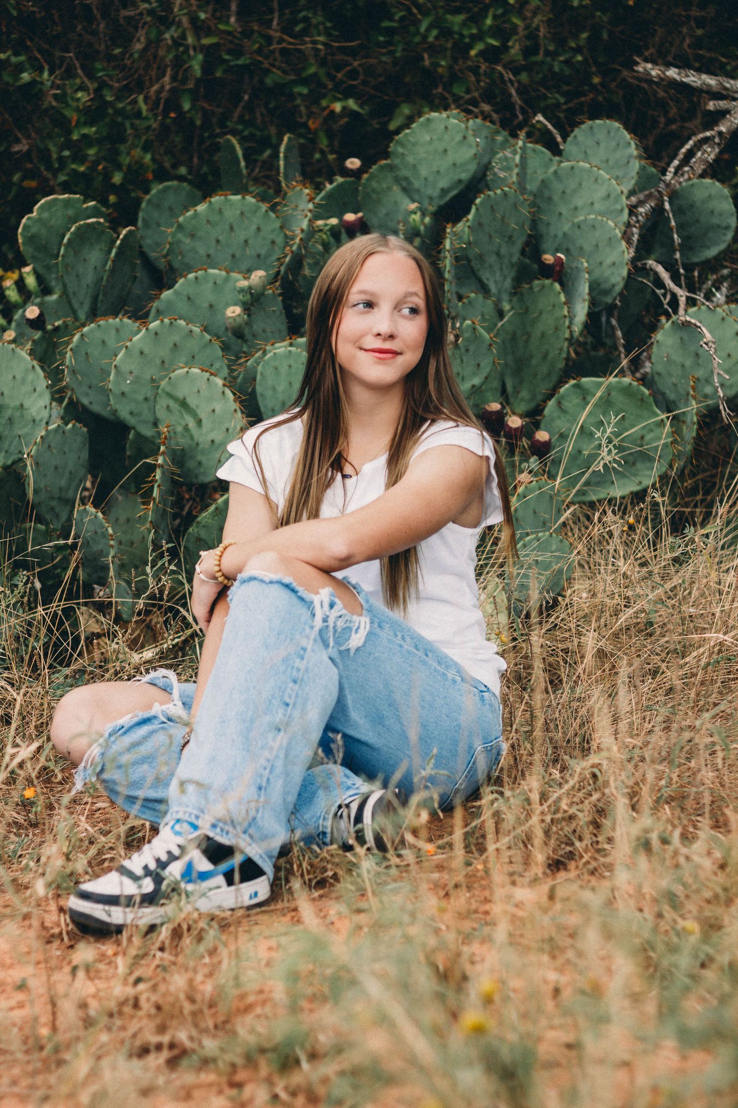 Senior girl  in white tee and ripped jeans sits cross-legged in dry grass before large prickly pear cactus senior session