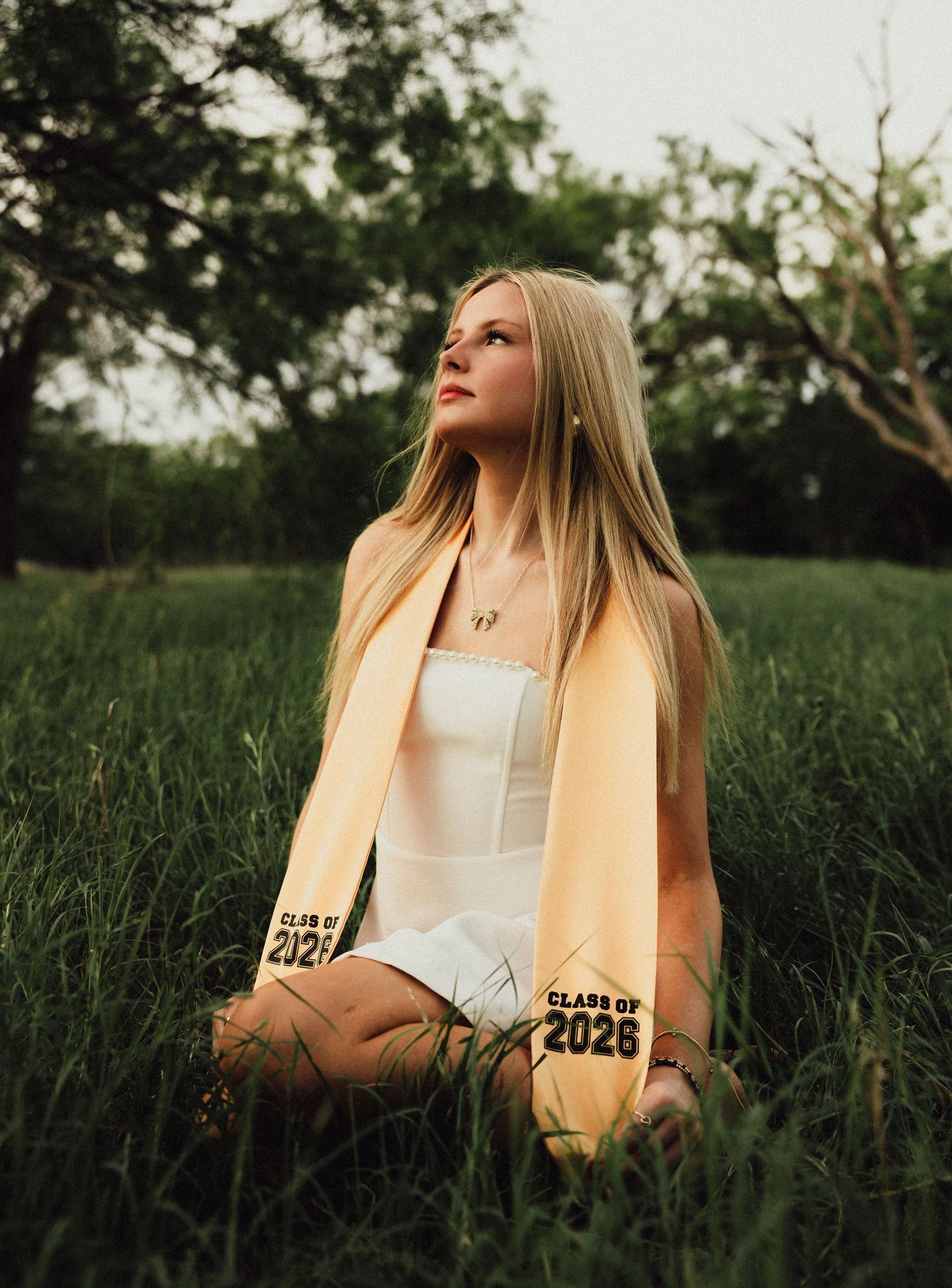 A blonde high school senior sits in lush green grass wearing a white dress and gold Class of 2026 graduation stole. Abilene, TX