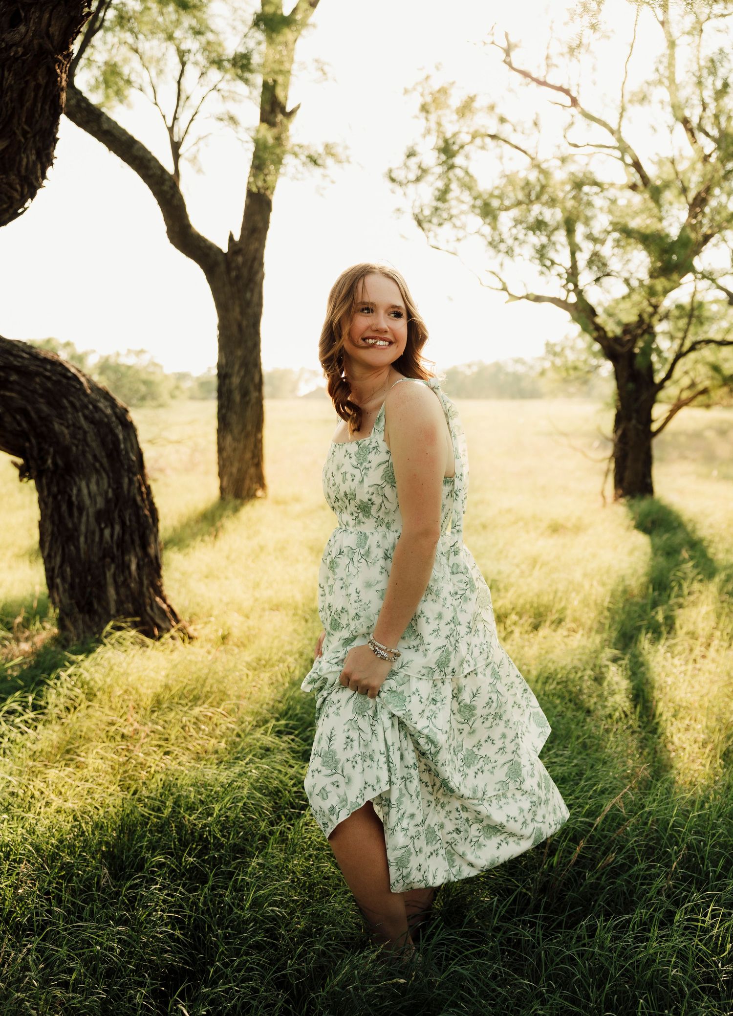 Abilene TX Senior in floral sundress smiling outdoors among trees in golden sunlight.