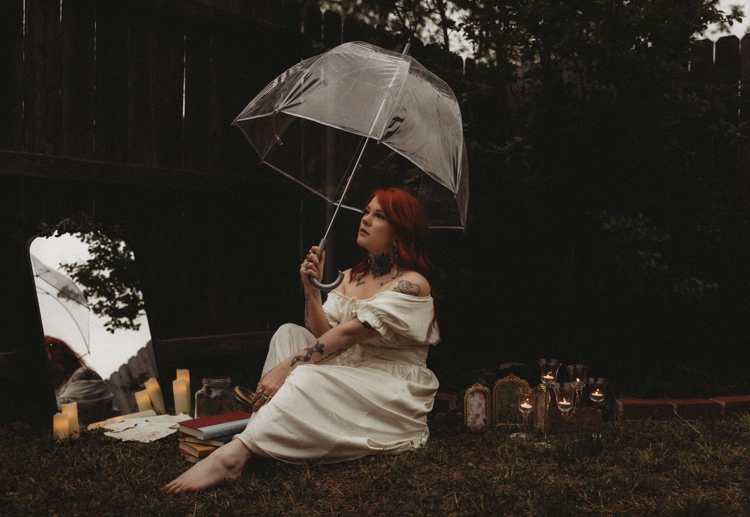 Woman in white dress sitting on grass holding a clear umbrella outdoors at dusk with candles nearby.