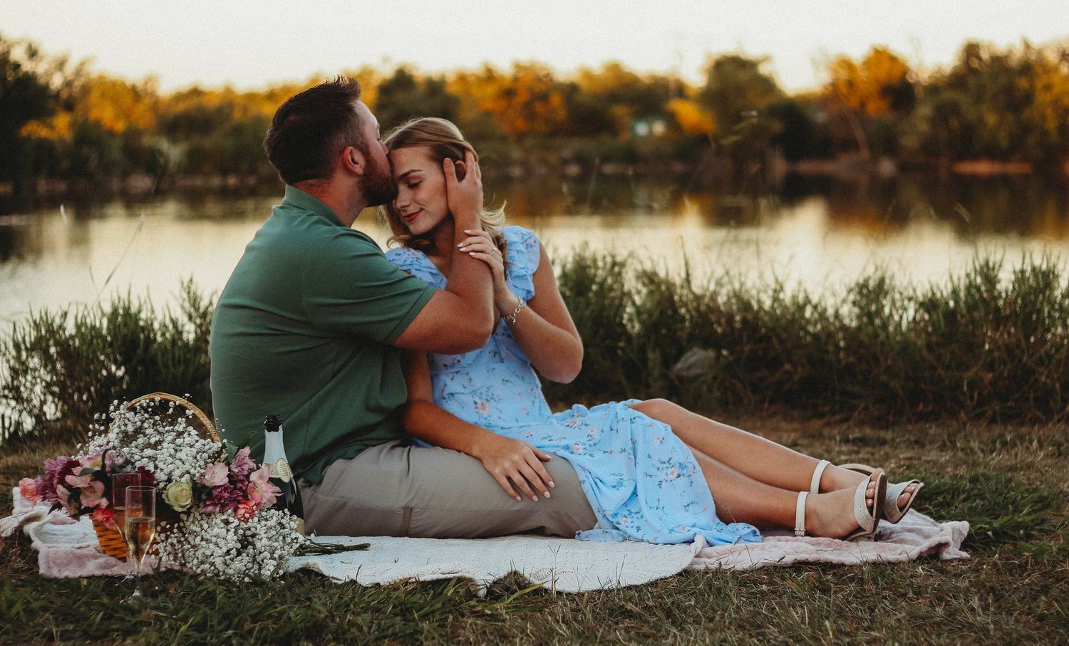 Engagement photoshoot couple sharing a romantic lakeside picnic on a blanket surrounded by flowers at sunset.