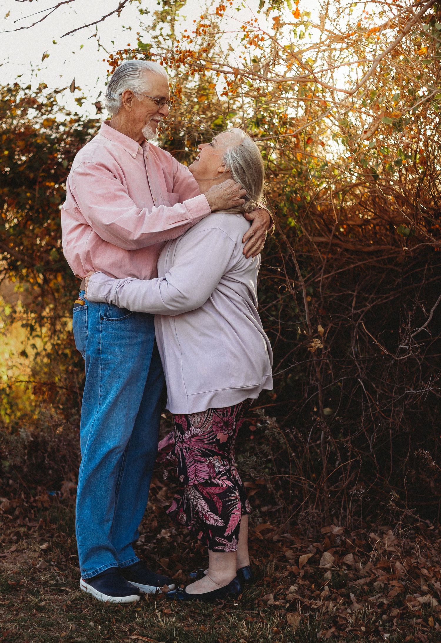 An elderly couple embraces and gazes lovingly at each other outdoors among autumn foliage in warm golden light.