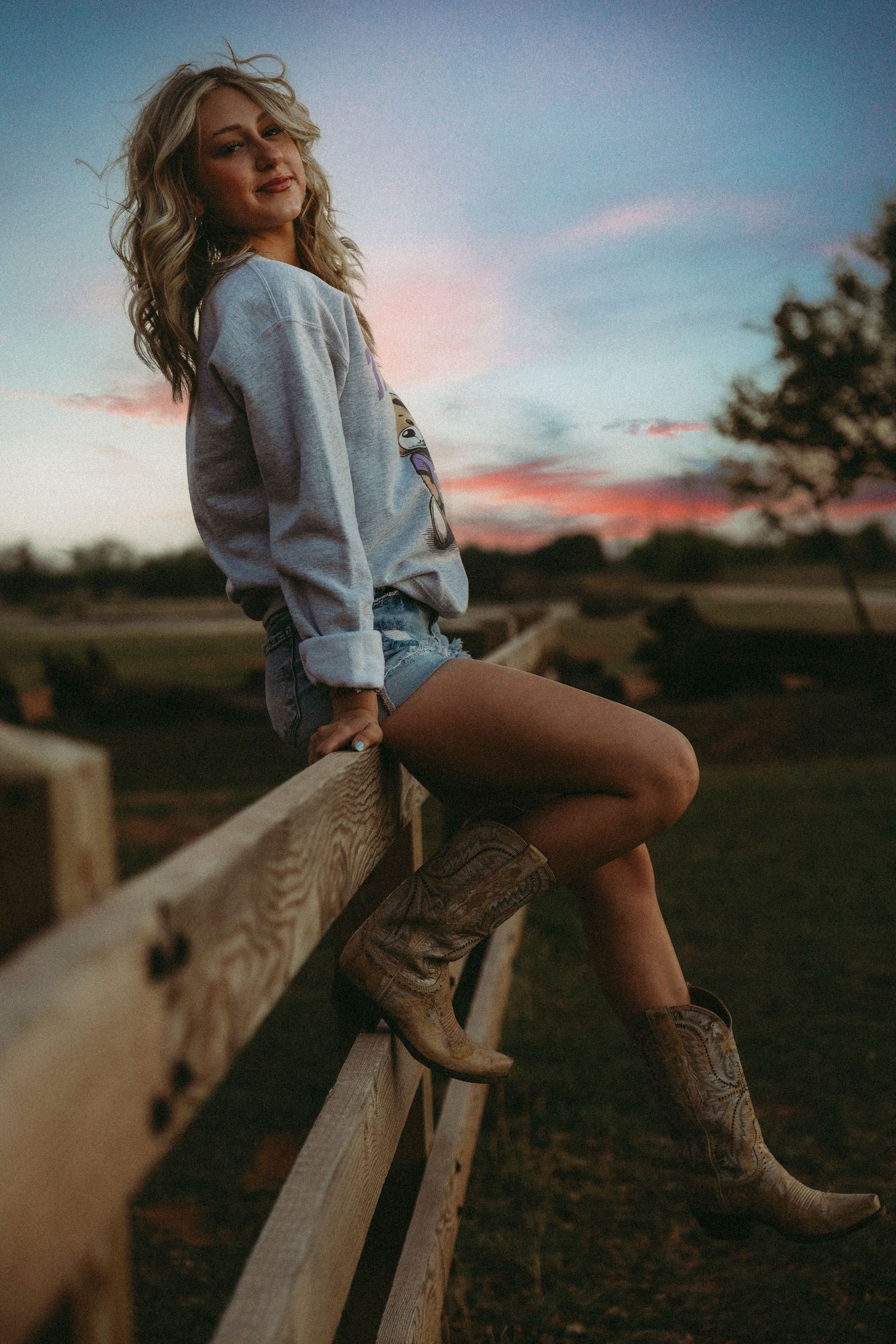 Senior girl in denim shorts and cowboy boots sitting on a wooden fence at sunset on a farm.