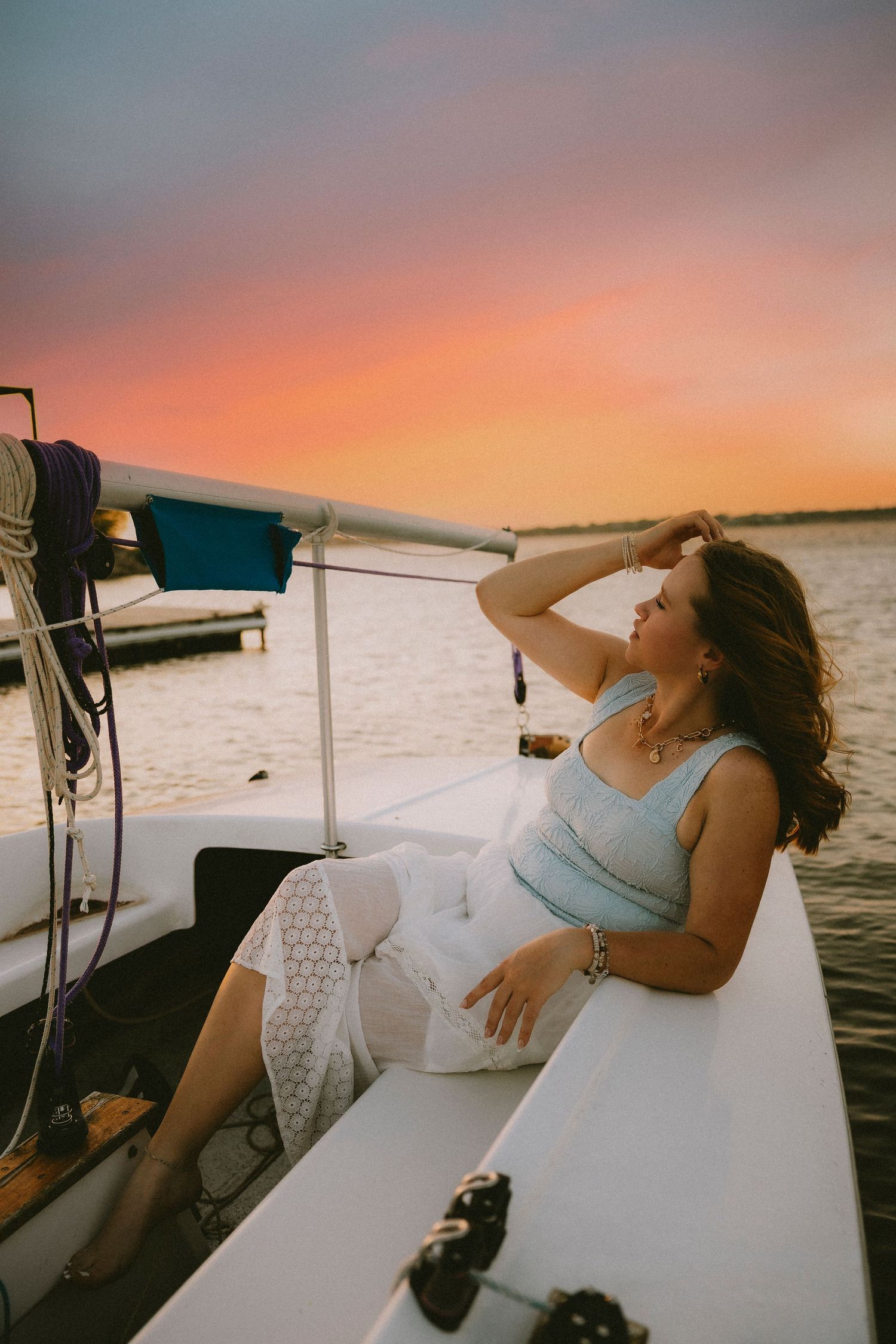 Abilene TX High Senior relaxing on a sailboat at sunset, wearing a blue top and white lace skirt, with a vibrant pink sky over the water.