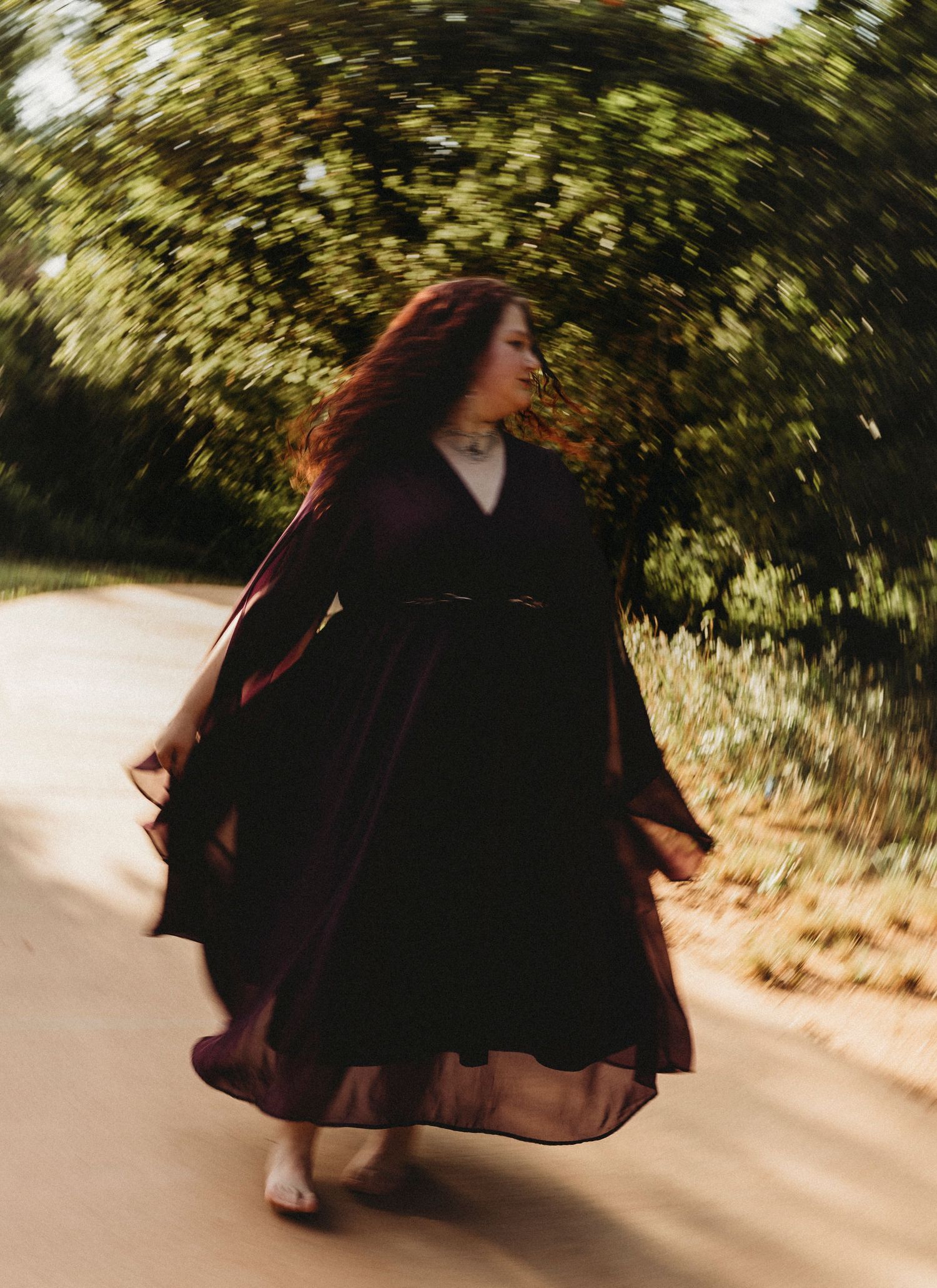 High school senior walking down a tree-lined road in a flowing burgundy dress during an outdoor senior portrait session in Texas.