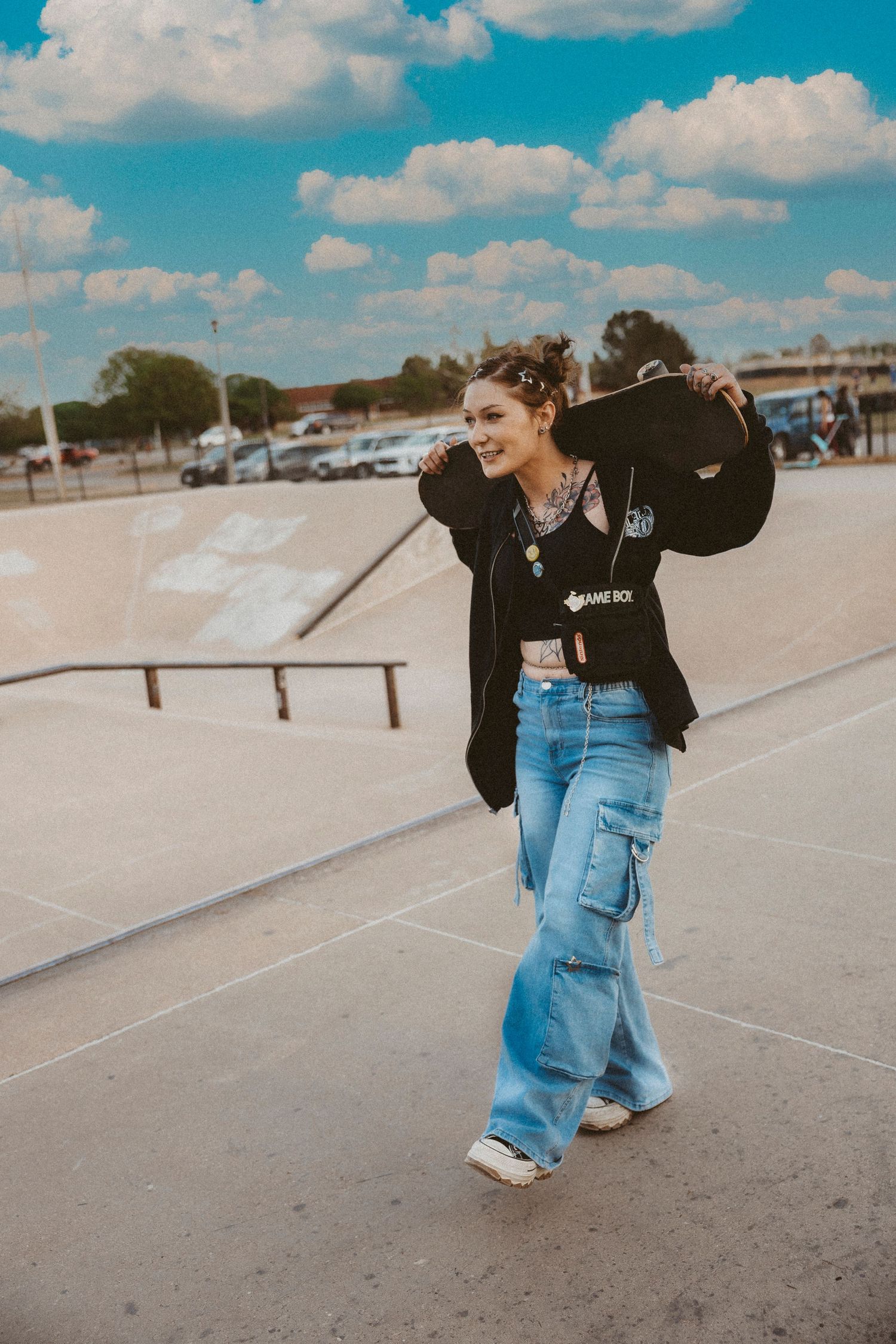 Young woman holding a skateboard at a Abilene TX skate park, wearing wide-leg jeans and a black hoodie under a blue sky.