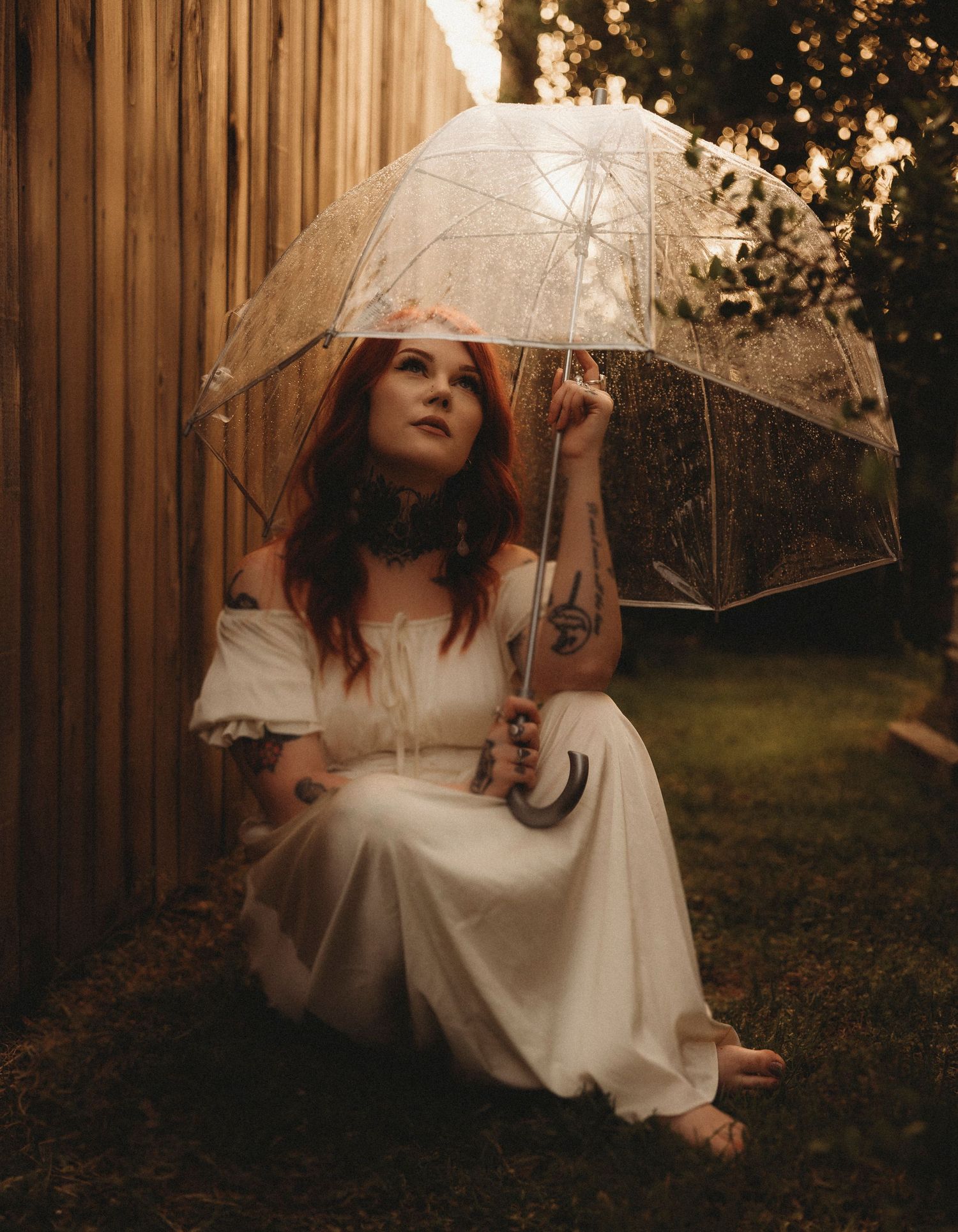 A redhead woman in a white dress sits barefoot outdoors holding a clear lace umbrella, with warm golden light behind her.