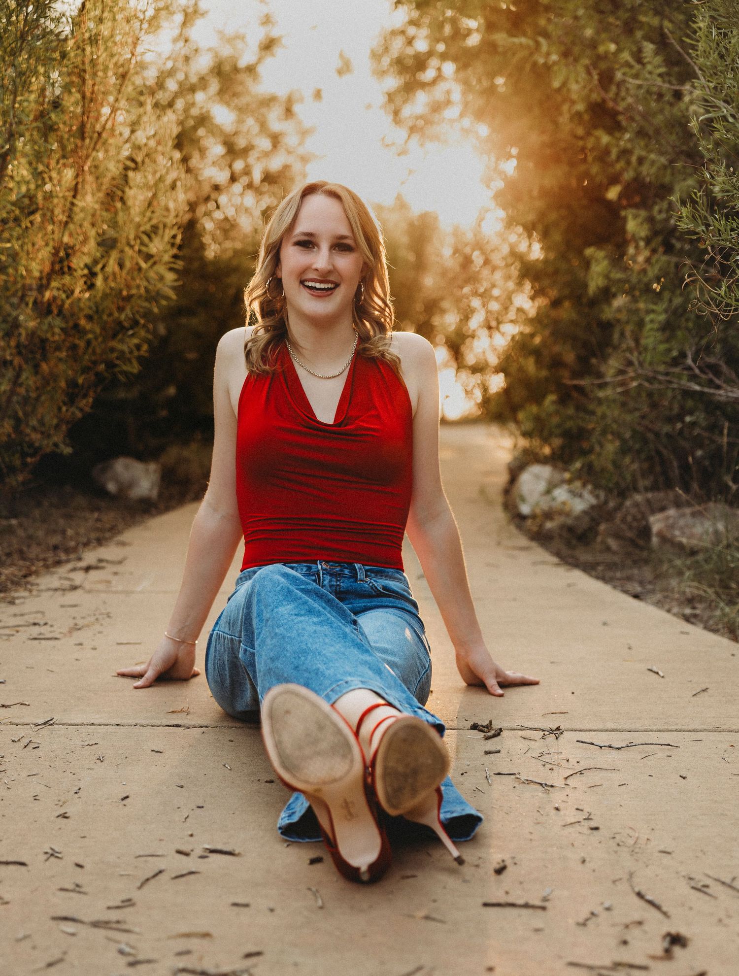 High school senior sitting on a tree-lined pathway in a red top and jeans during a golden hour senior portrait session in Abilene, Texas.