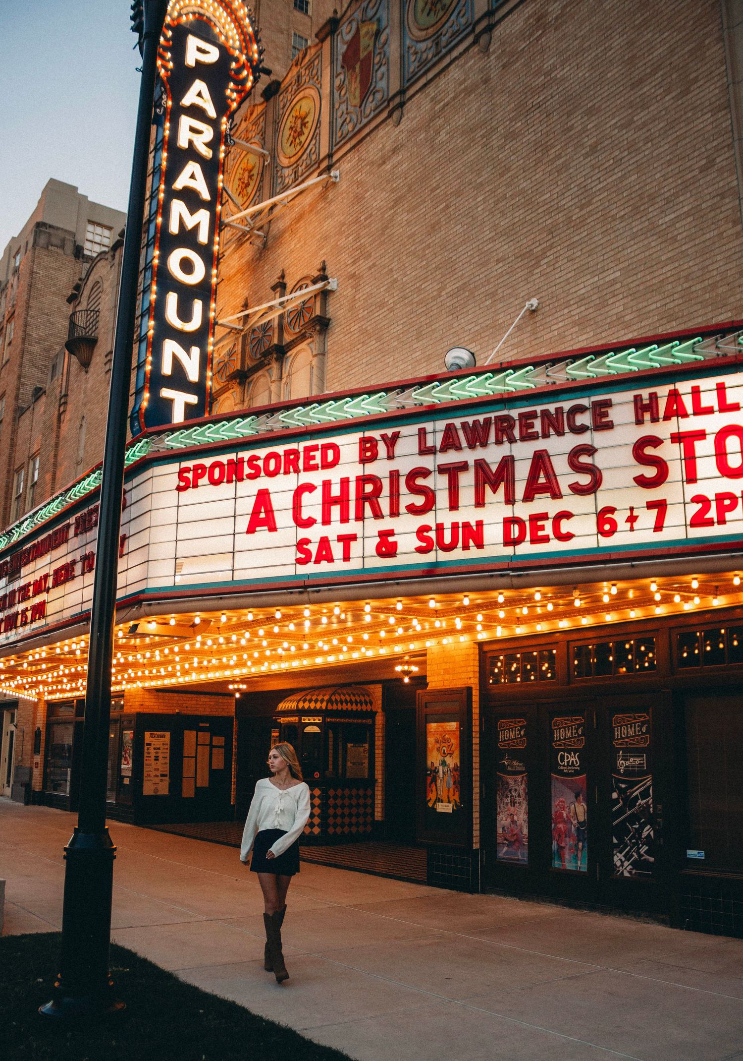 Vintage illuminated theater marquee signs on historic brick buildings with Christmas show advertisements at dusk.