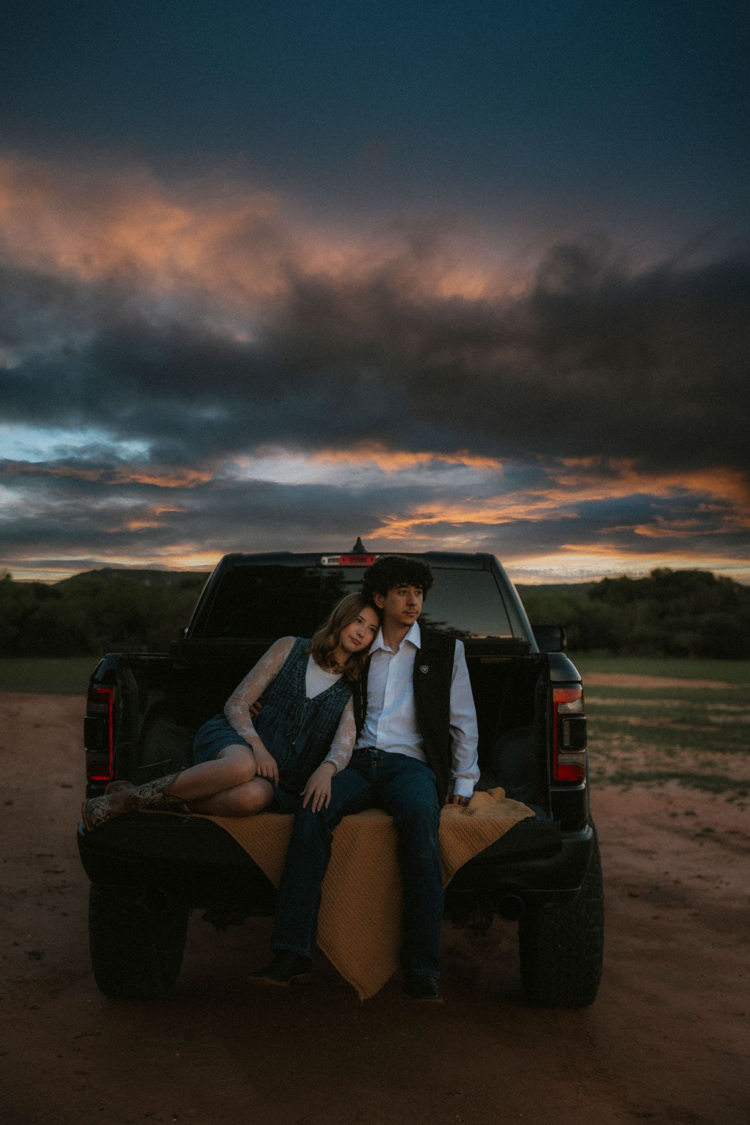 Couple sitting on truck tailgate at sunset with dramatic Texas cloudy sky in rural landscape.