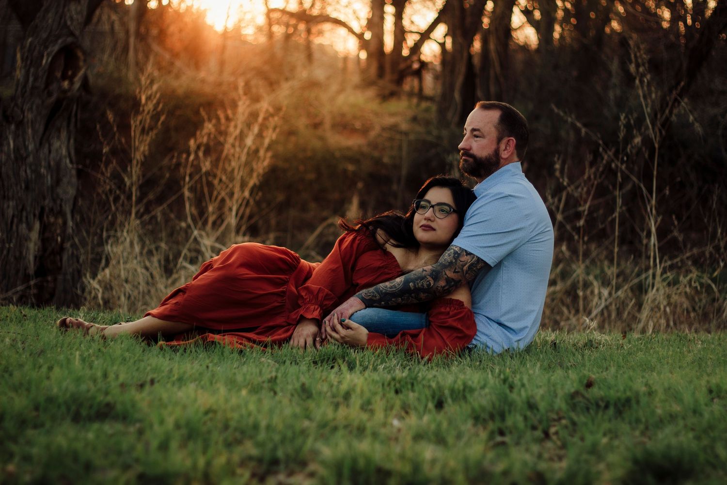 Couple sitting together at golden hour in a field, woman in red dress leaning on man with tattoos.