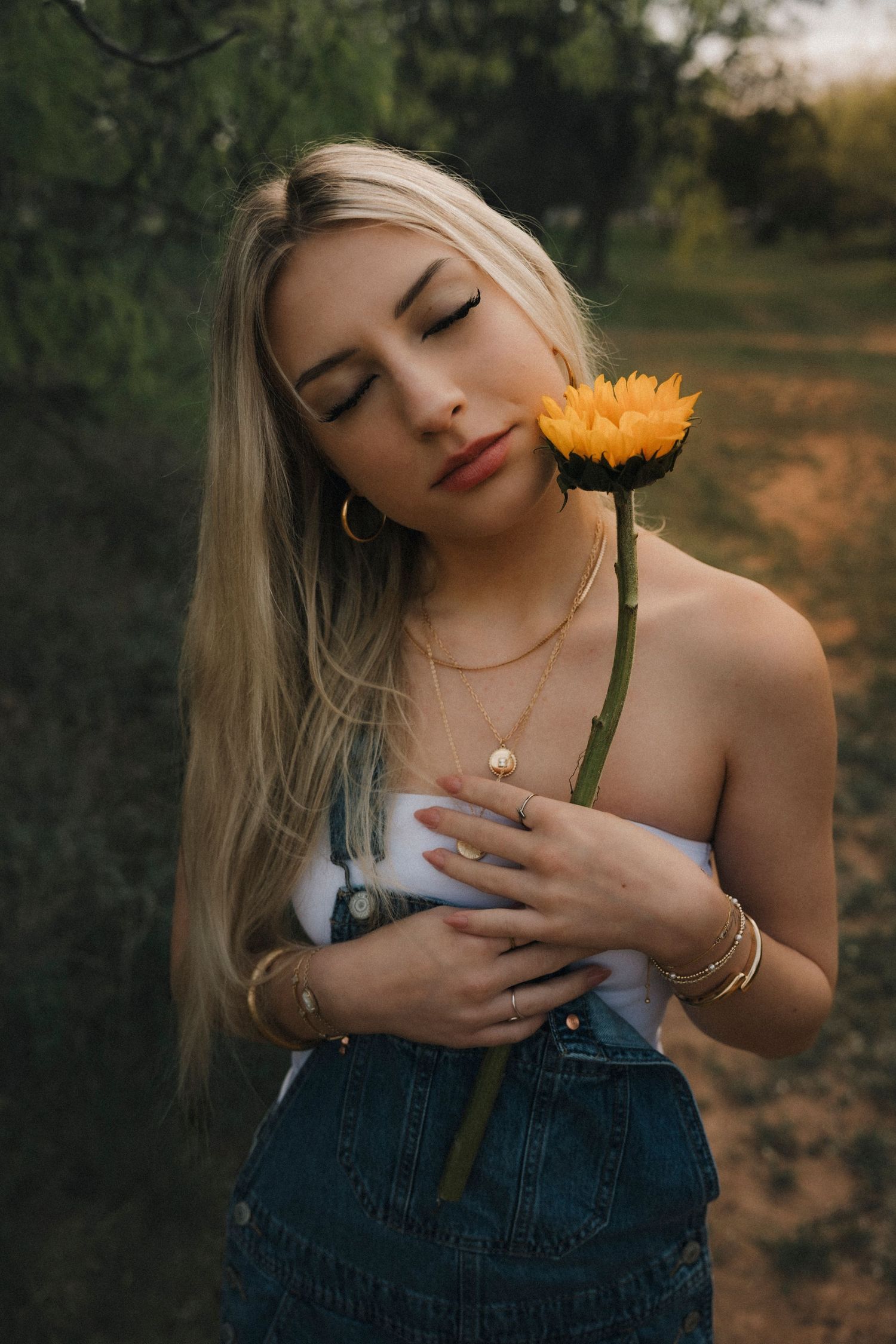 Senior girl photoshoot denim overalls holding a yellow sunflower with eyes closed outdoors. 90s style edit