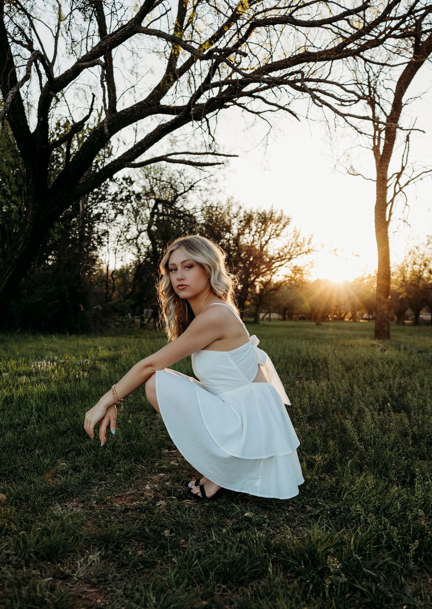 Wylie High School Senior in white dress crouching in grassy field at golden sunset with bare trees in background.