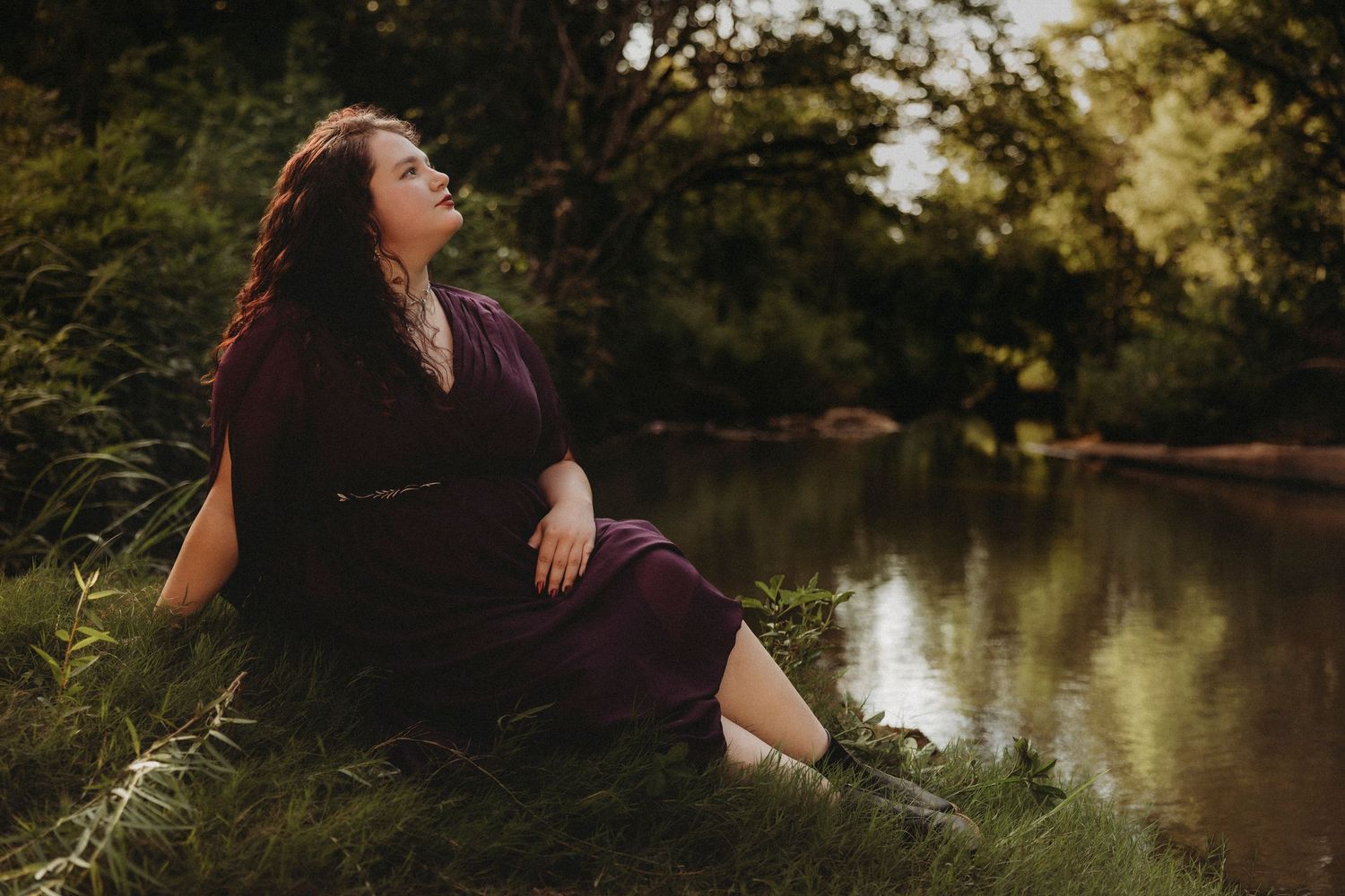 Golden hour senior photos of a high school senior in a burgundy dress sitting beside a creek during an outdoor senior portrait session in Ab