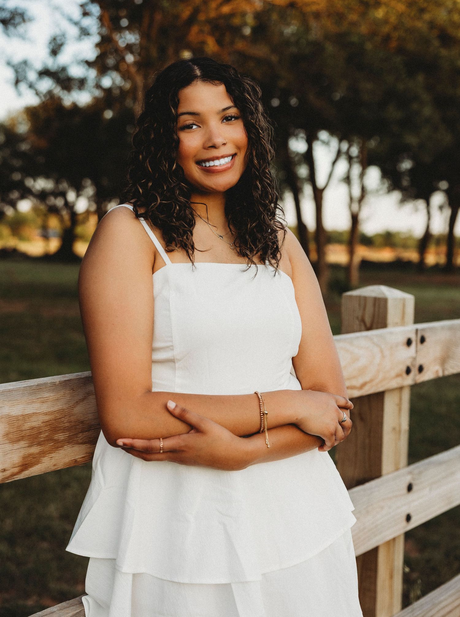 Abilene High School Senior in white dress smiling outdoors by wooden fence with trees in golden hour light.
