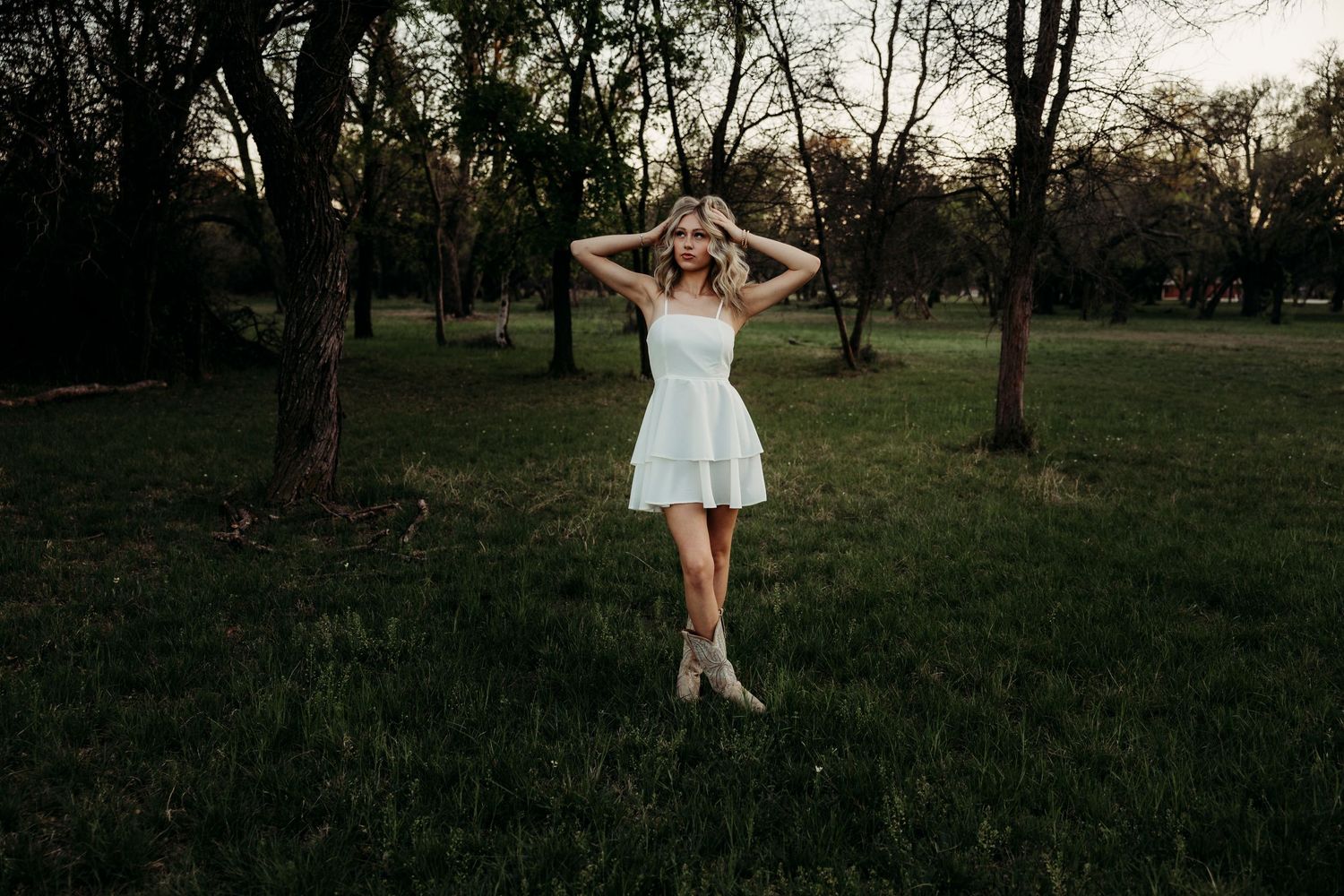Senior photoshoot in a white mini dress and boots poses in a lush green park surrounded by trees.