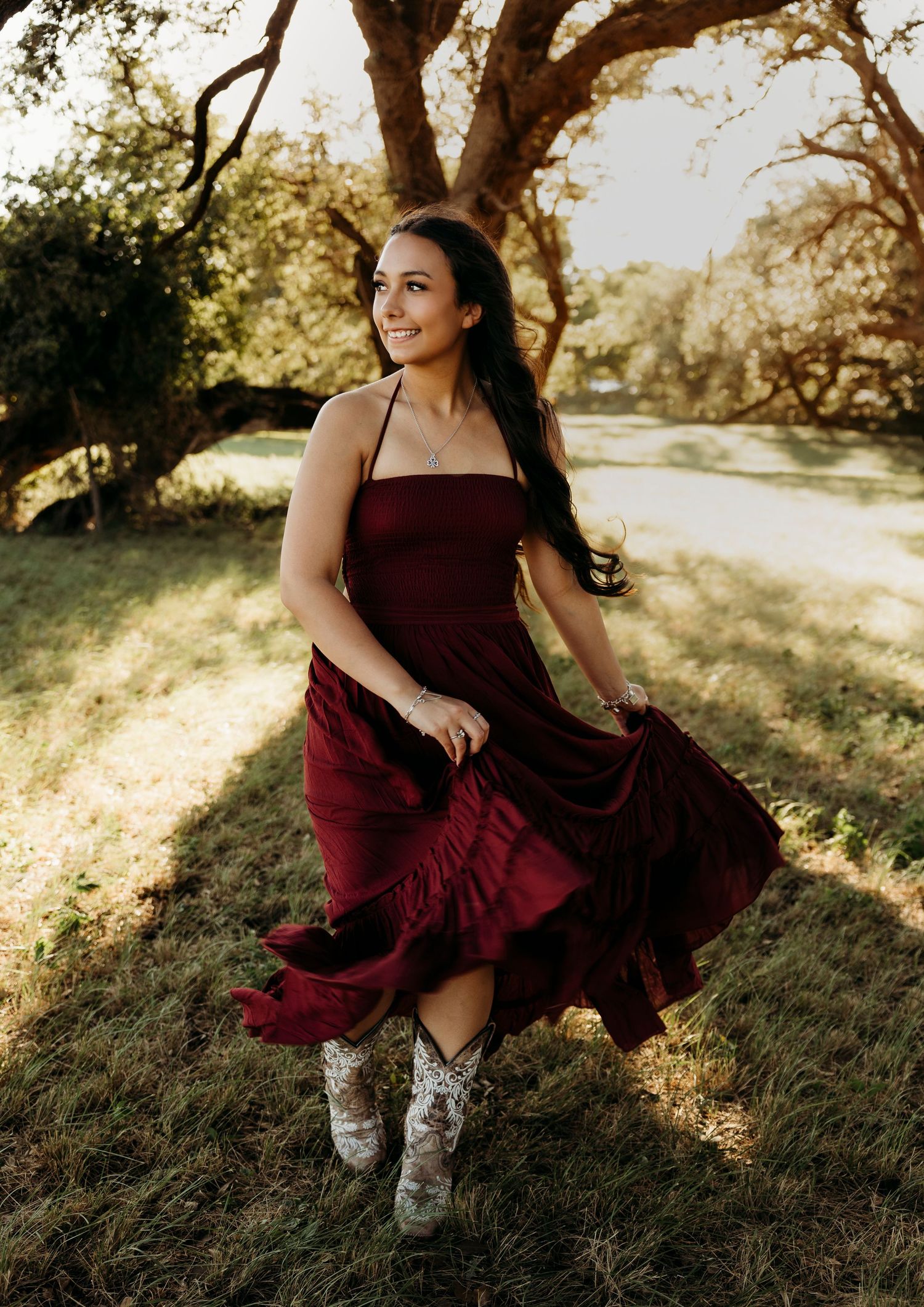 Elegant senior photos of a high school senior twirling in a burgundy dress beneath oak trees during an outdoor senior portrait session in Ab