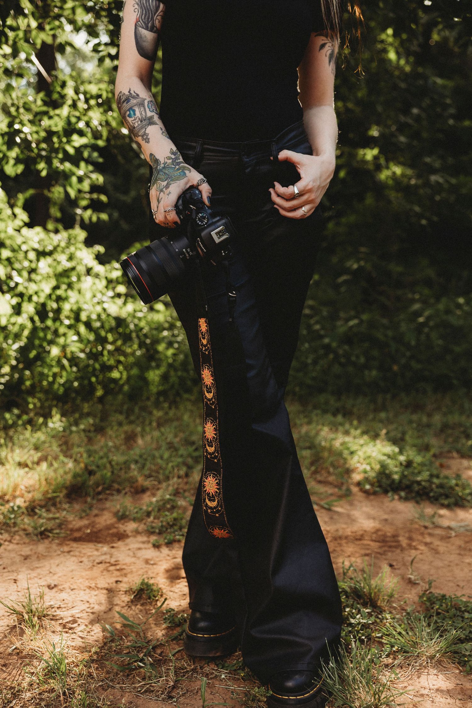 Tattooed photographer holding a camera wearing black flare pants with sun embroidery outdoors. Abilene Texas