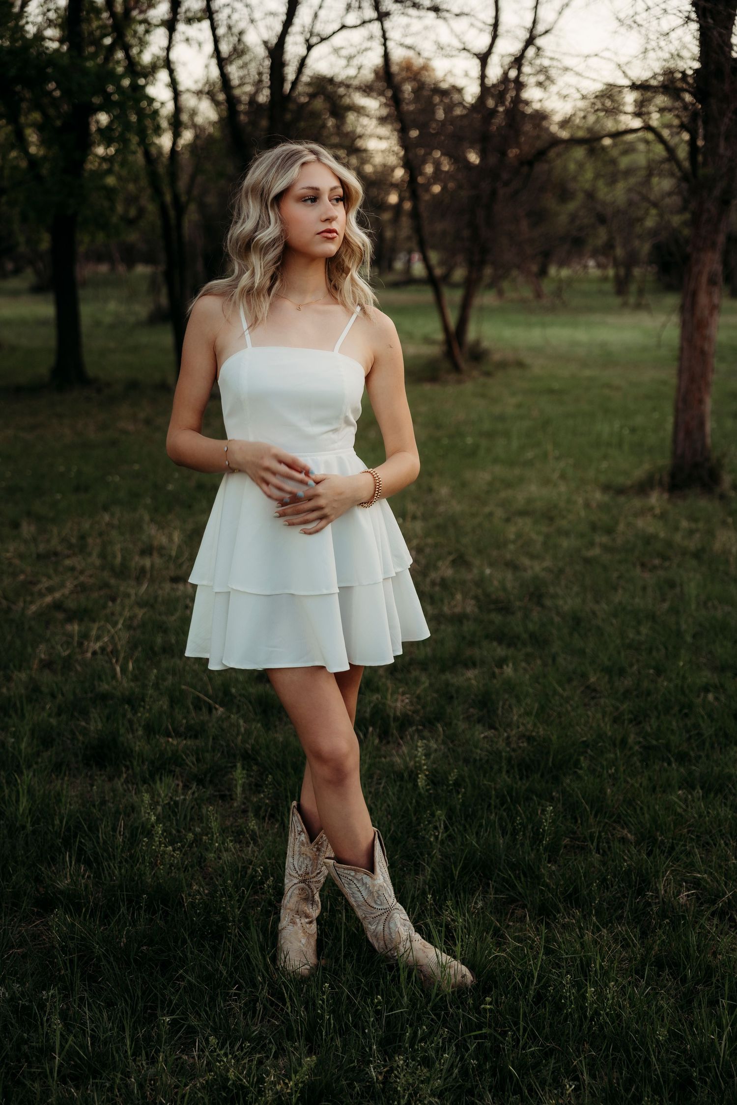 Wylie High School Senior in white mini dress and cowboy boots standing in a lush green field at dusk.