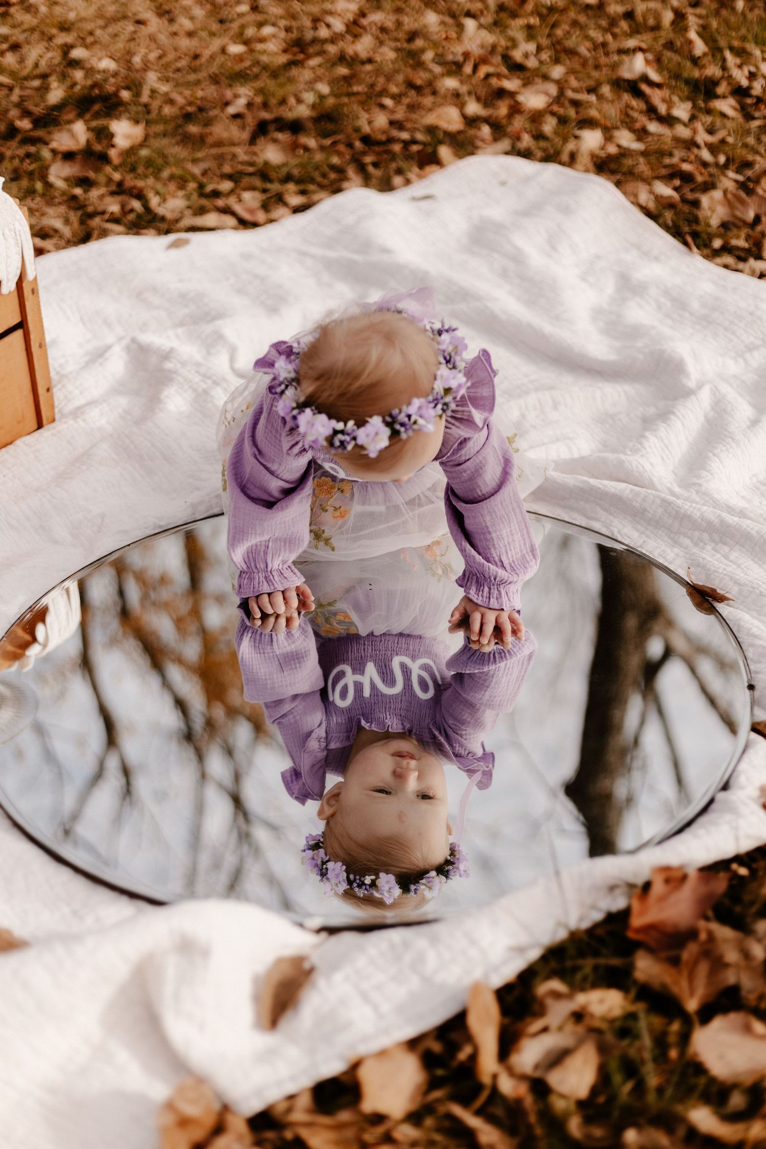 Baby in purple outfit lying on white blanket surrounded by autumn leaves.