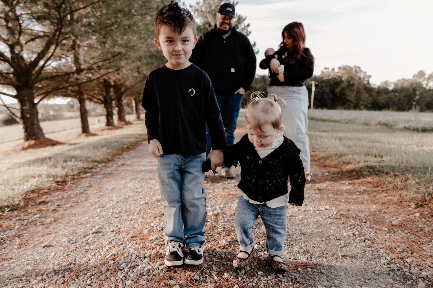 Two young children walking together on a gravel path with adults in background.