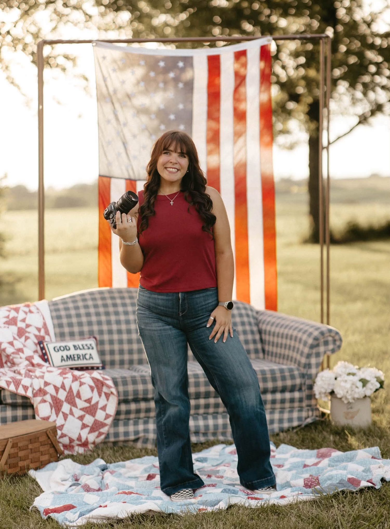 Woman holding camera stands on quilt before American flag backdrop outdoors in red top and jeans.
