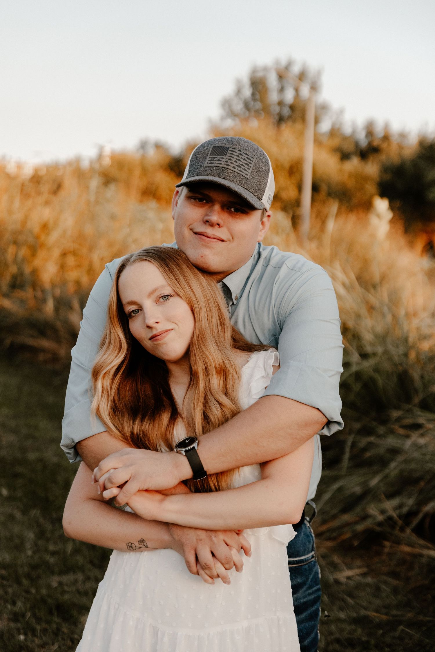 A romantic couple embraces in a golden sunset field wearing casual attire.