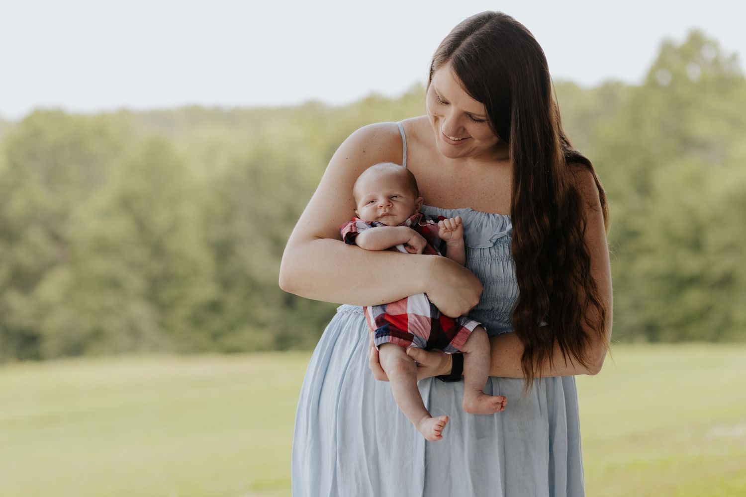 A young mother in a light blue dress tenderly holds her newborn baby outdoors in a lush green field.