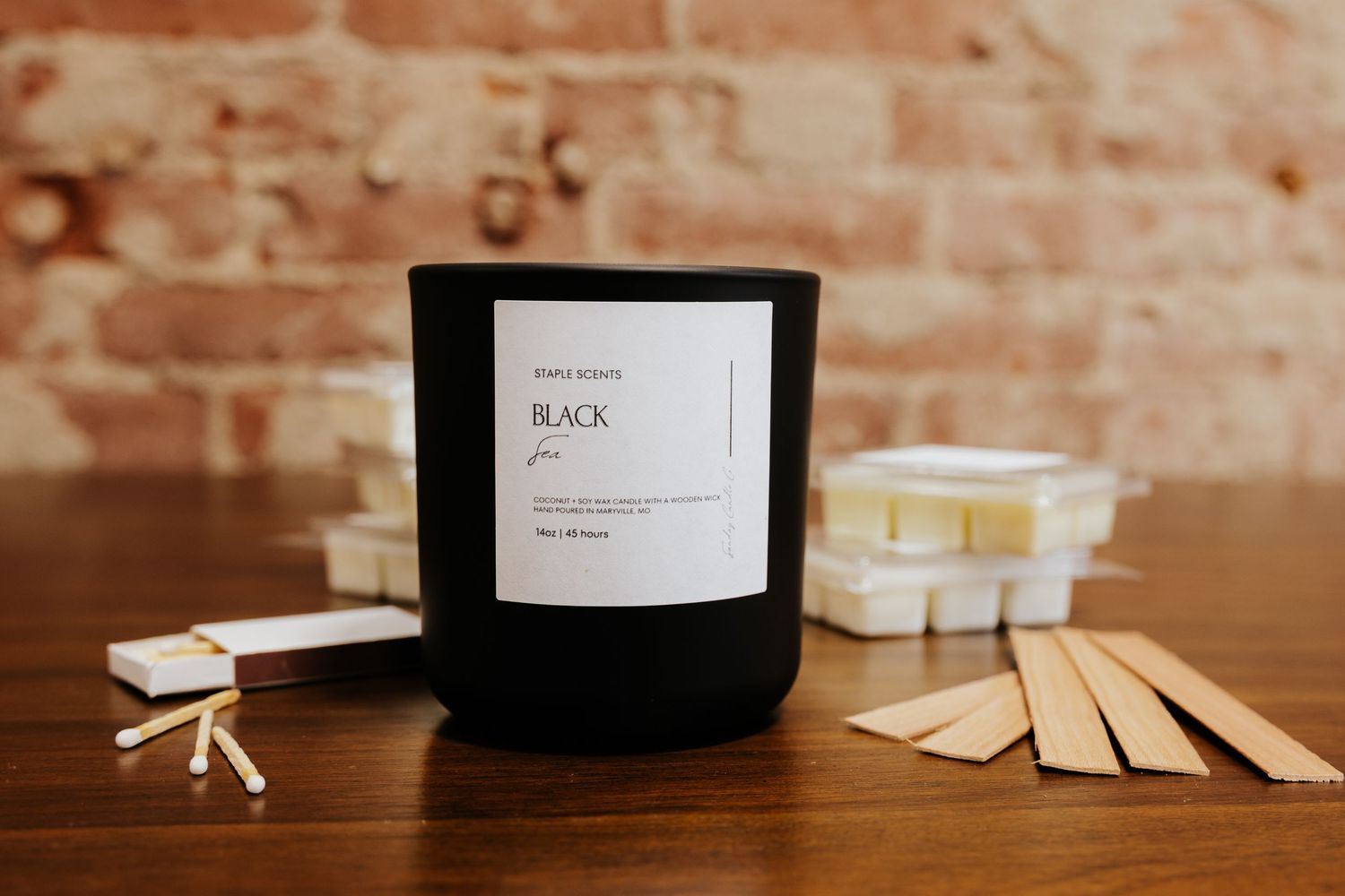 Black scented candle in matte jar with matches and wax melts on wooden table against brick wall.