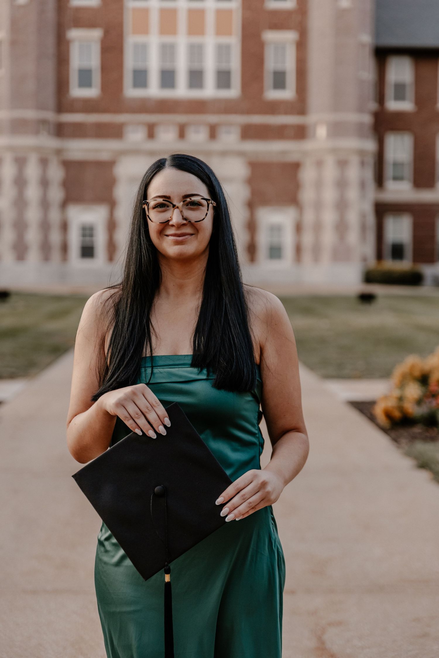 Graduate holding cap in emerald green dress outside brick building.