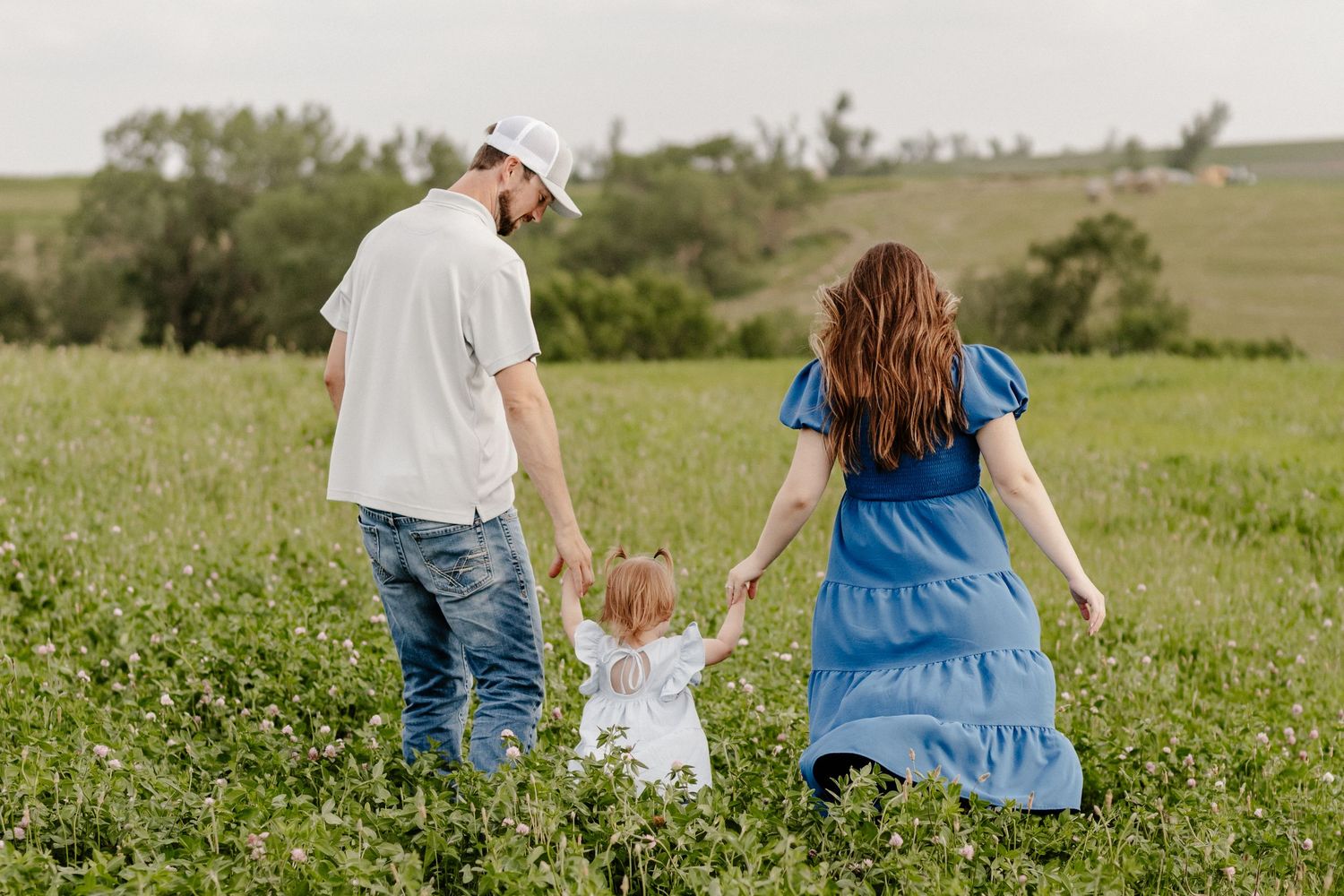 A family of three walks hand-in-hand through a lush green field, parents guiding their young child.