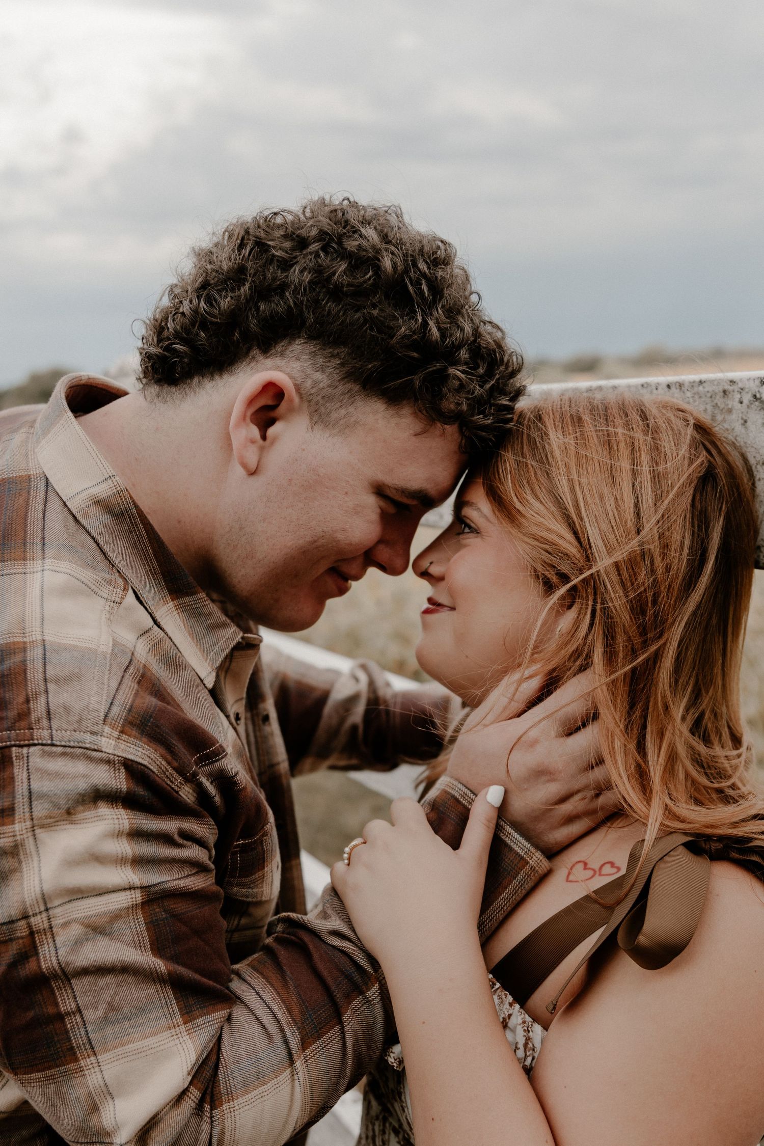 A couple shares a romantic forehead-to-forehead moment outdoors, the man in a plaid shirt tenderly holding the woman's face.