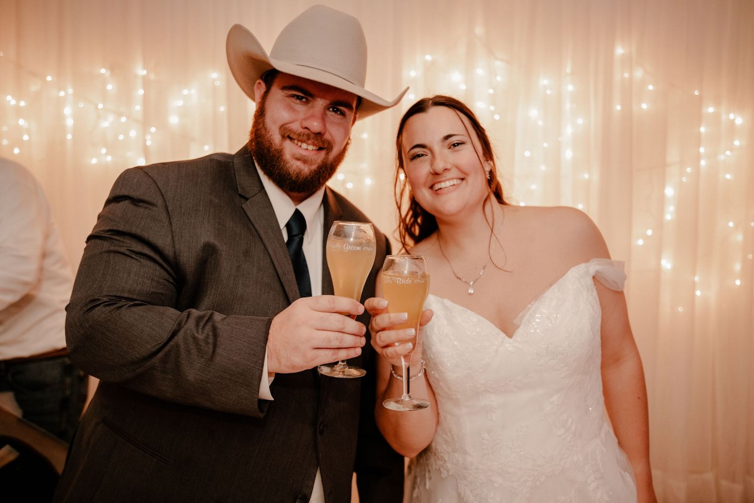 Couple toasts with champagne glasses at wedding reception with string lights backdrop.