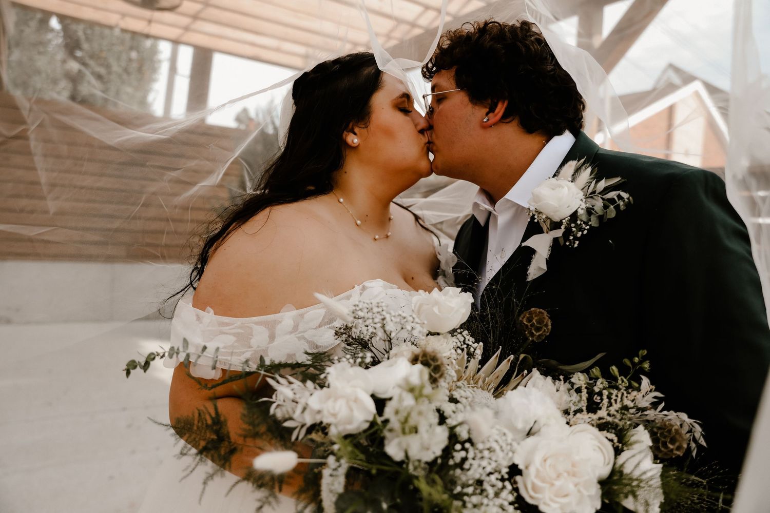 A romantic wedding moment captured with a couple kissing while holding a white floral bouquet in a light-filled venue.