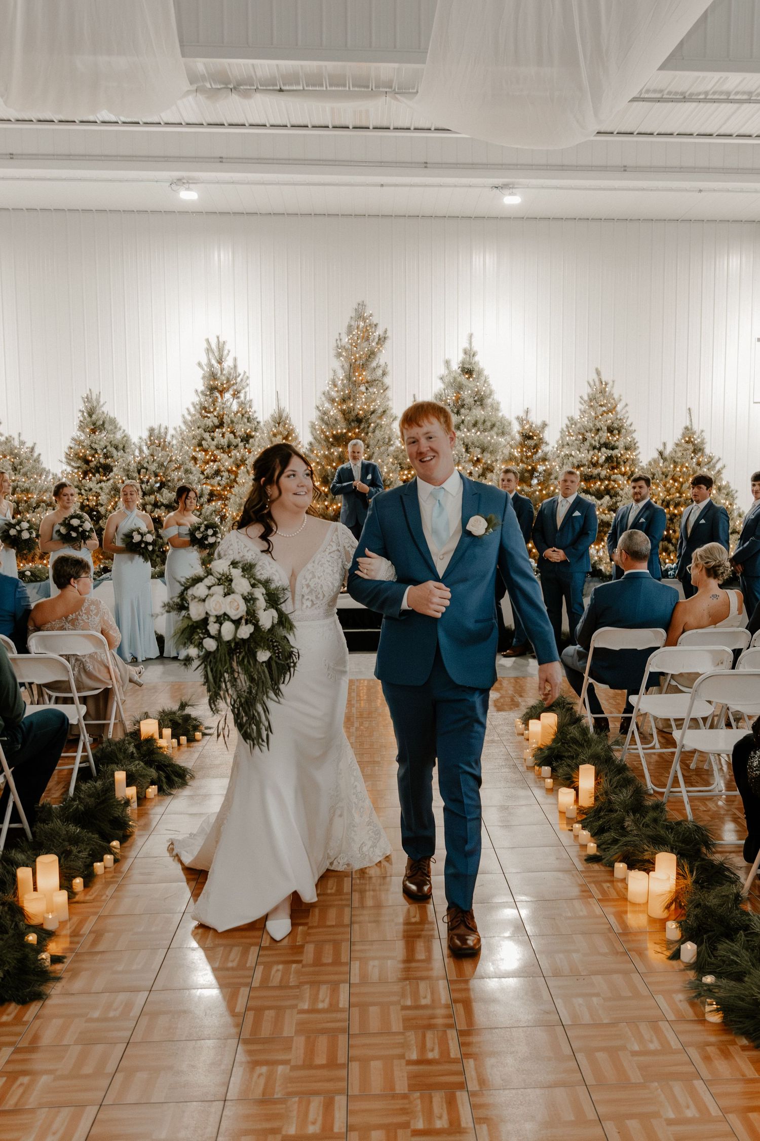 Couple walks down aisle at wedding reception with lit candles and gold Christmas trees lining the path.