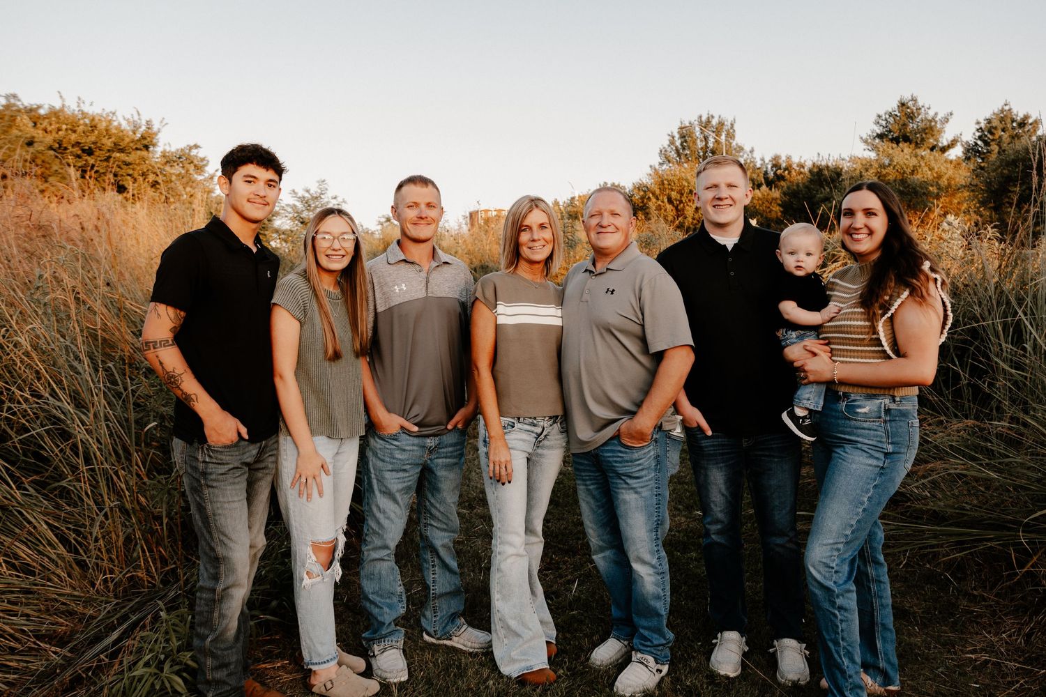 Casual outdoor family portrait of eight people standing together in tall grass at sunset.