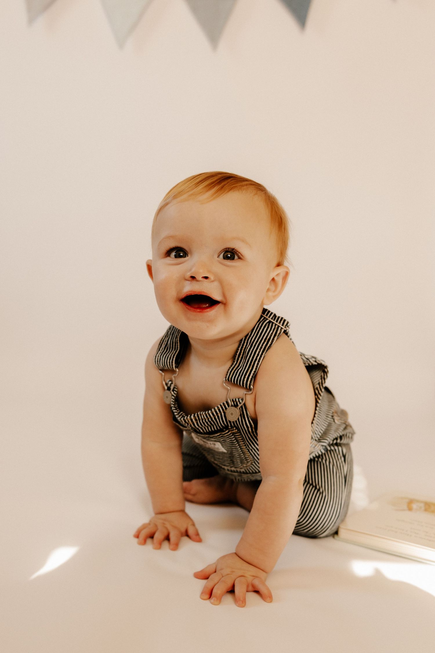 Smiling baby in striped denim overalls sitting on white floor with bunting banner decorations in background.