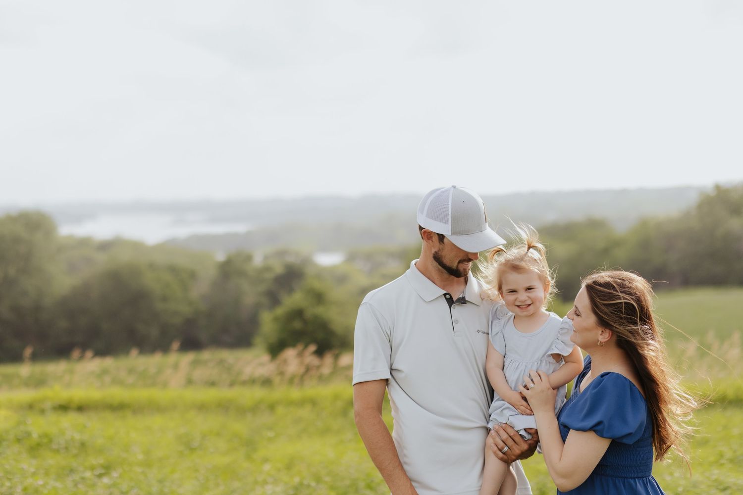 A happy young family with a toddler poses in a green meadow with a scenic water view in the background.